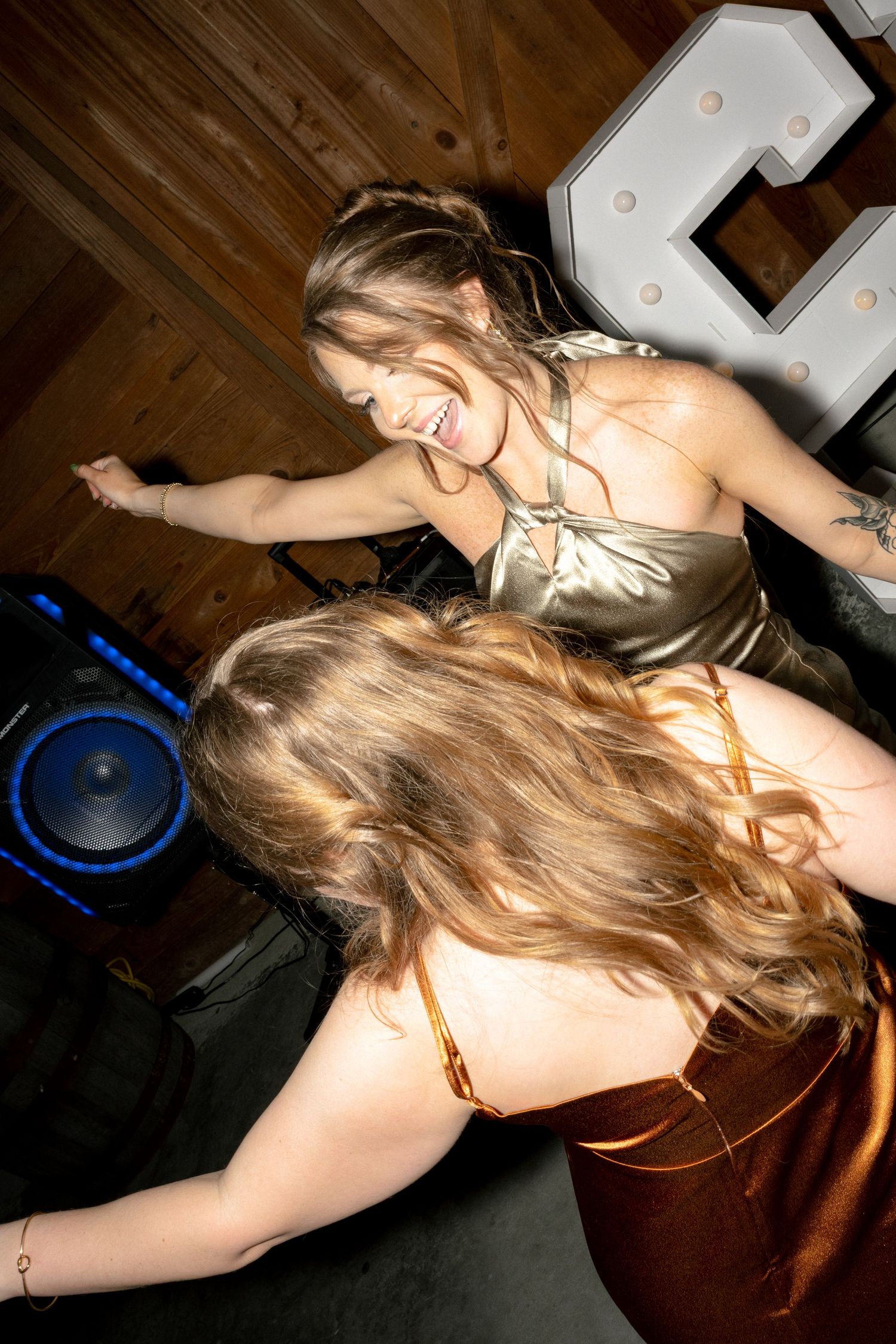 Two women in satin dresses dance near a DJ speaker at a rustic barn wedding reception.