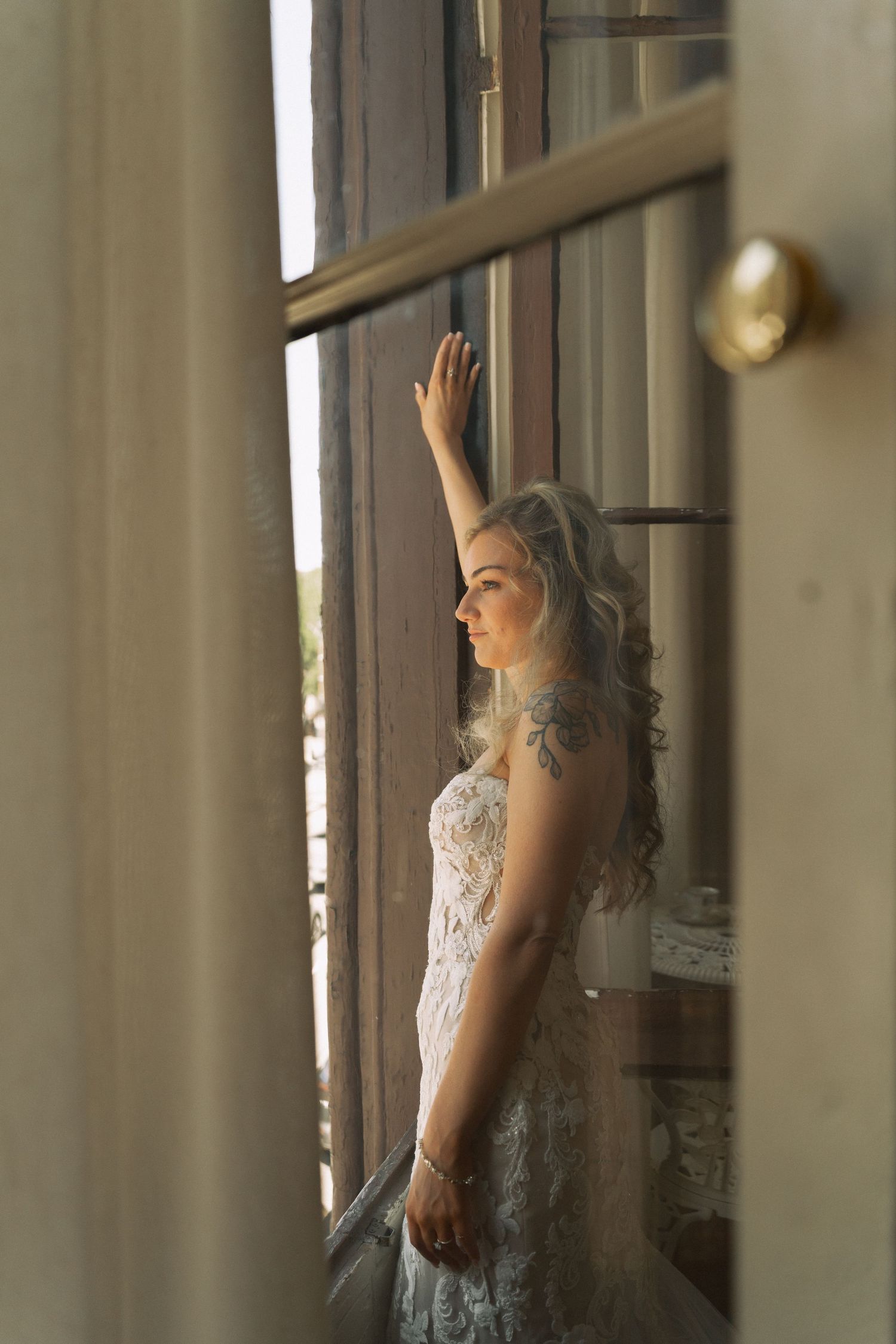 Blonde bride in lace wedding dress with tattoos poses by sunlit window, hand raised against glass pane.