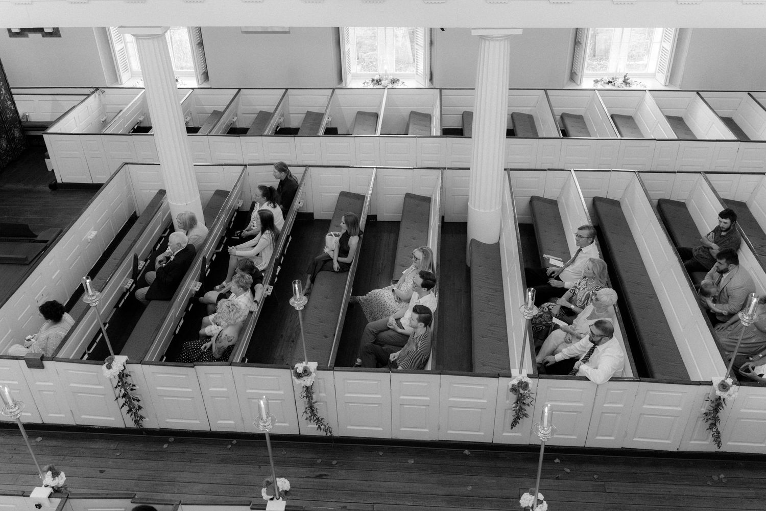 Aerial black and white view of wedding guests seated in white church pews decorated with greenery garlands.