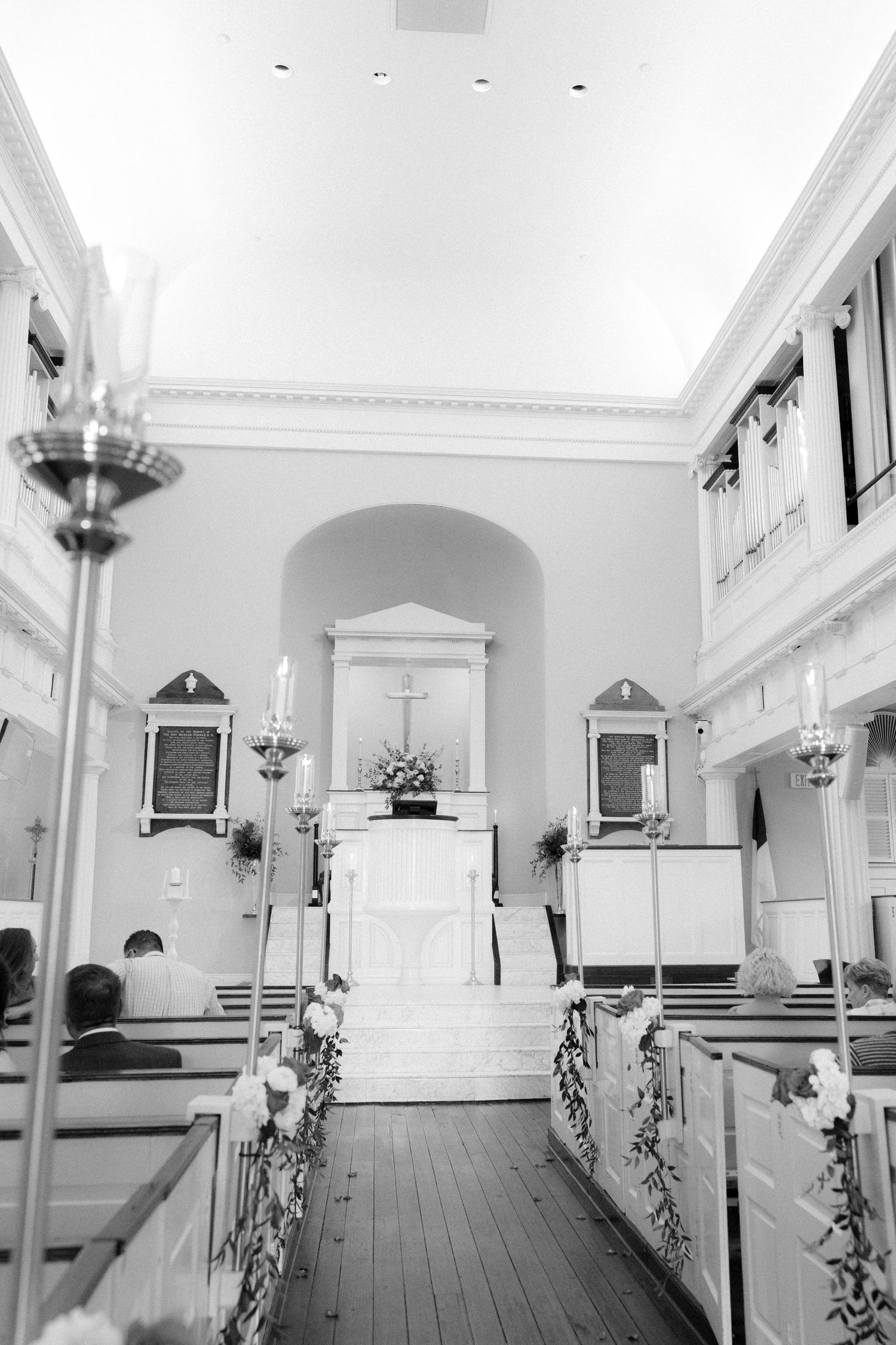 Black and white interior of a decorated church with pews, candles, and floral arrangements for a wedding ceremony.