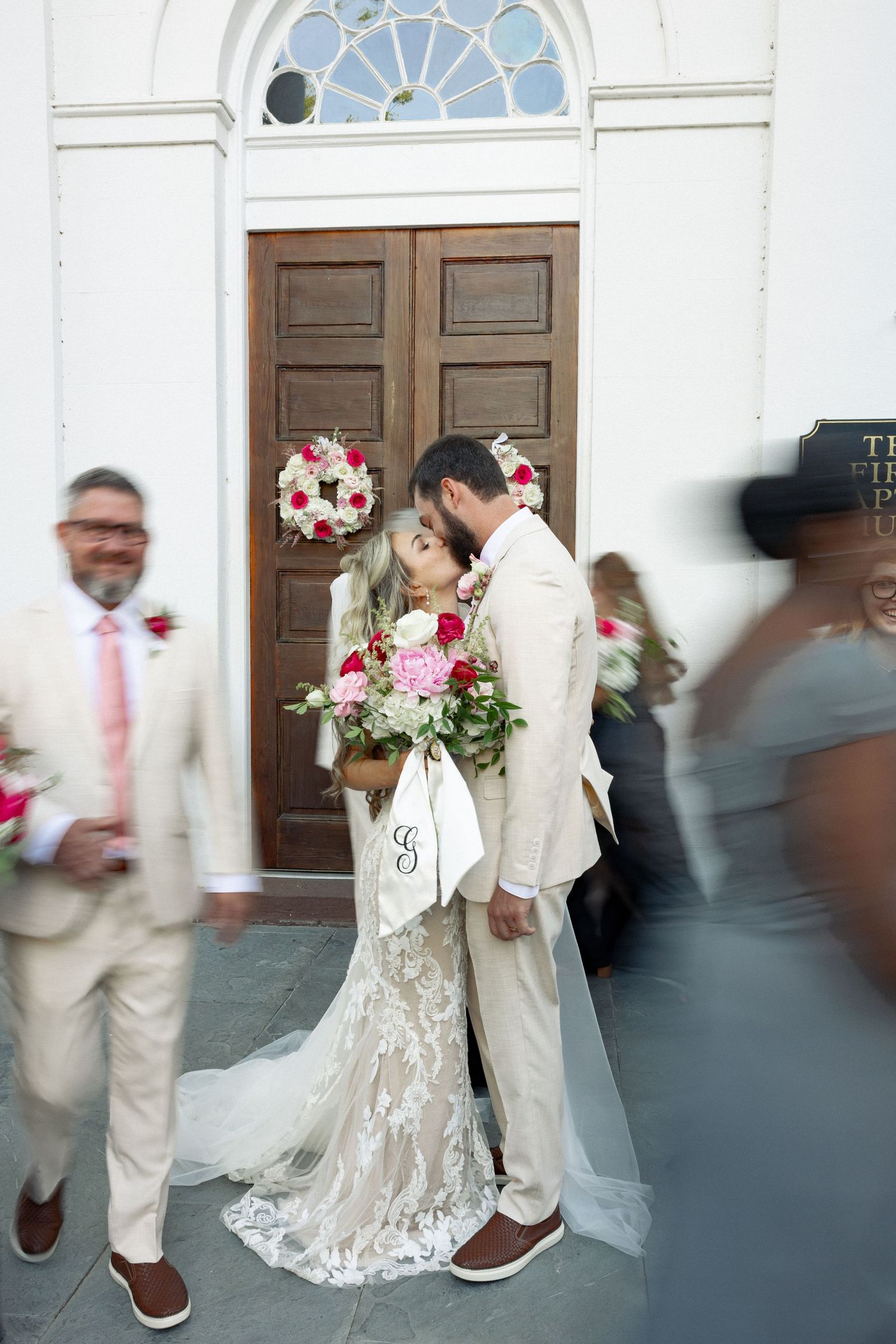 Bride and groom sharing a kiss outside First Baptist Church, bride holding pink and red floral bouquet.