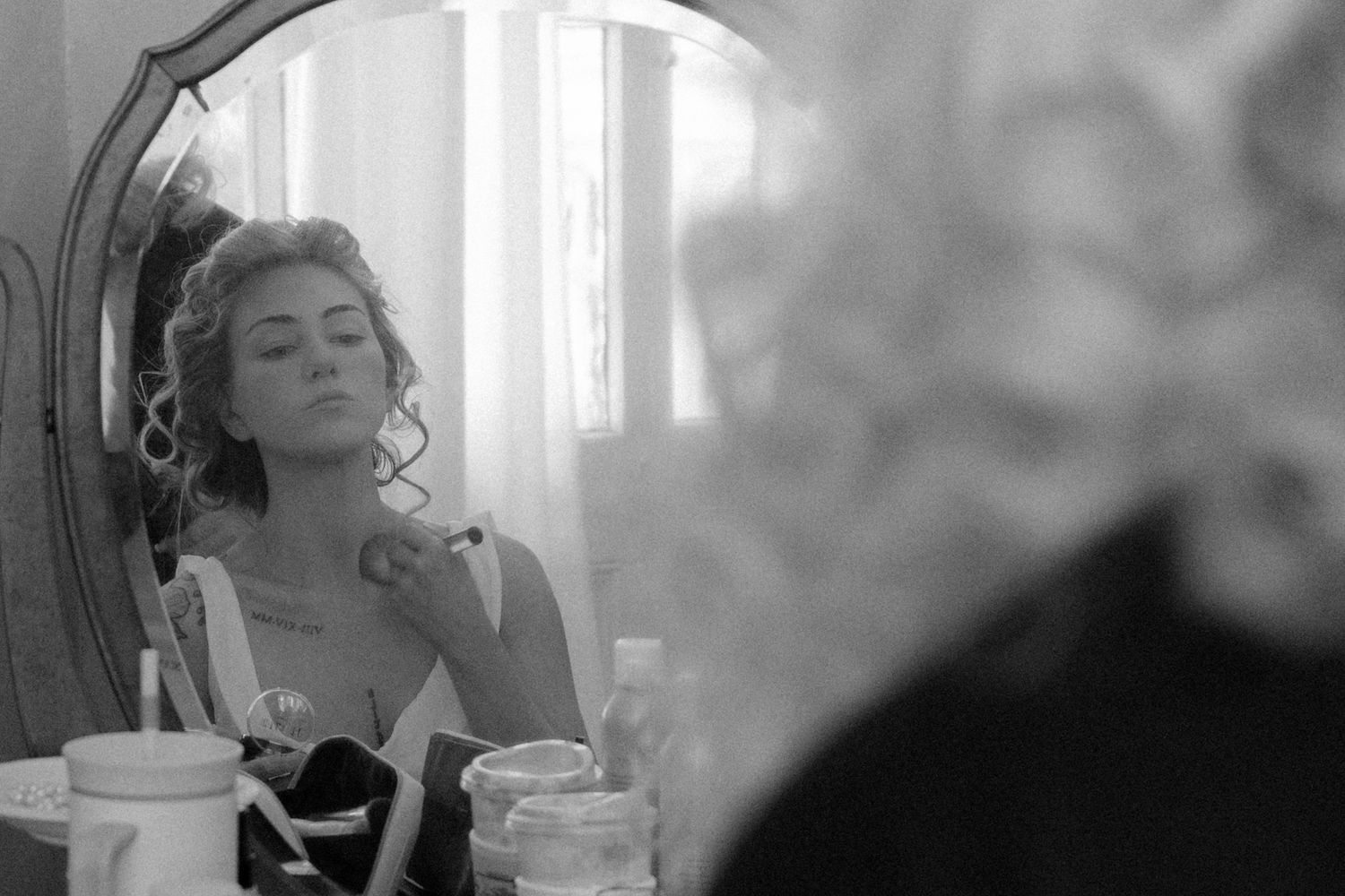 Black and white photo of a young woman applying makeup at a vintage vanity mirror in an elegant room.