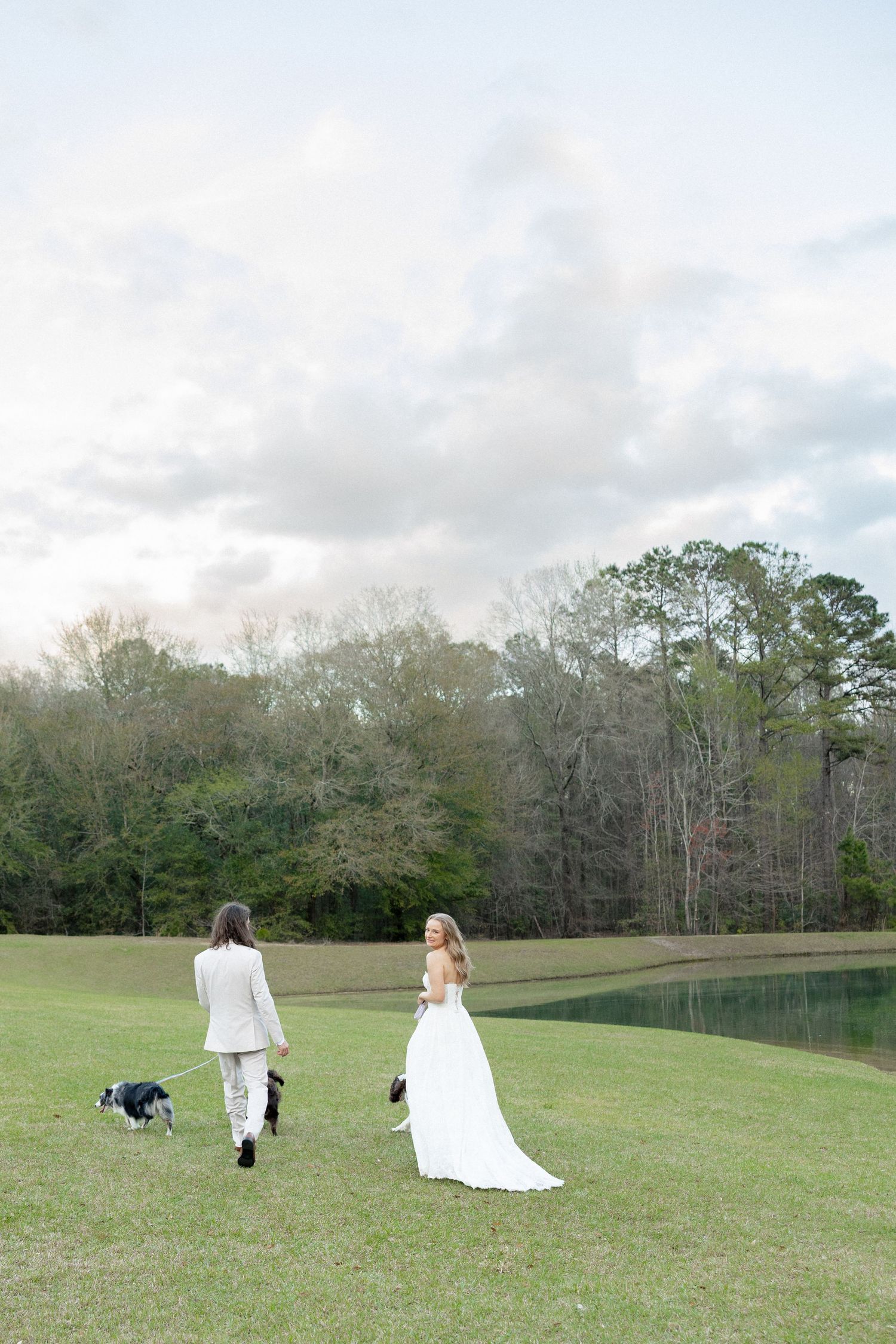 Bride and groom walking dog by a pond in lush green field on their wedding day.