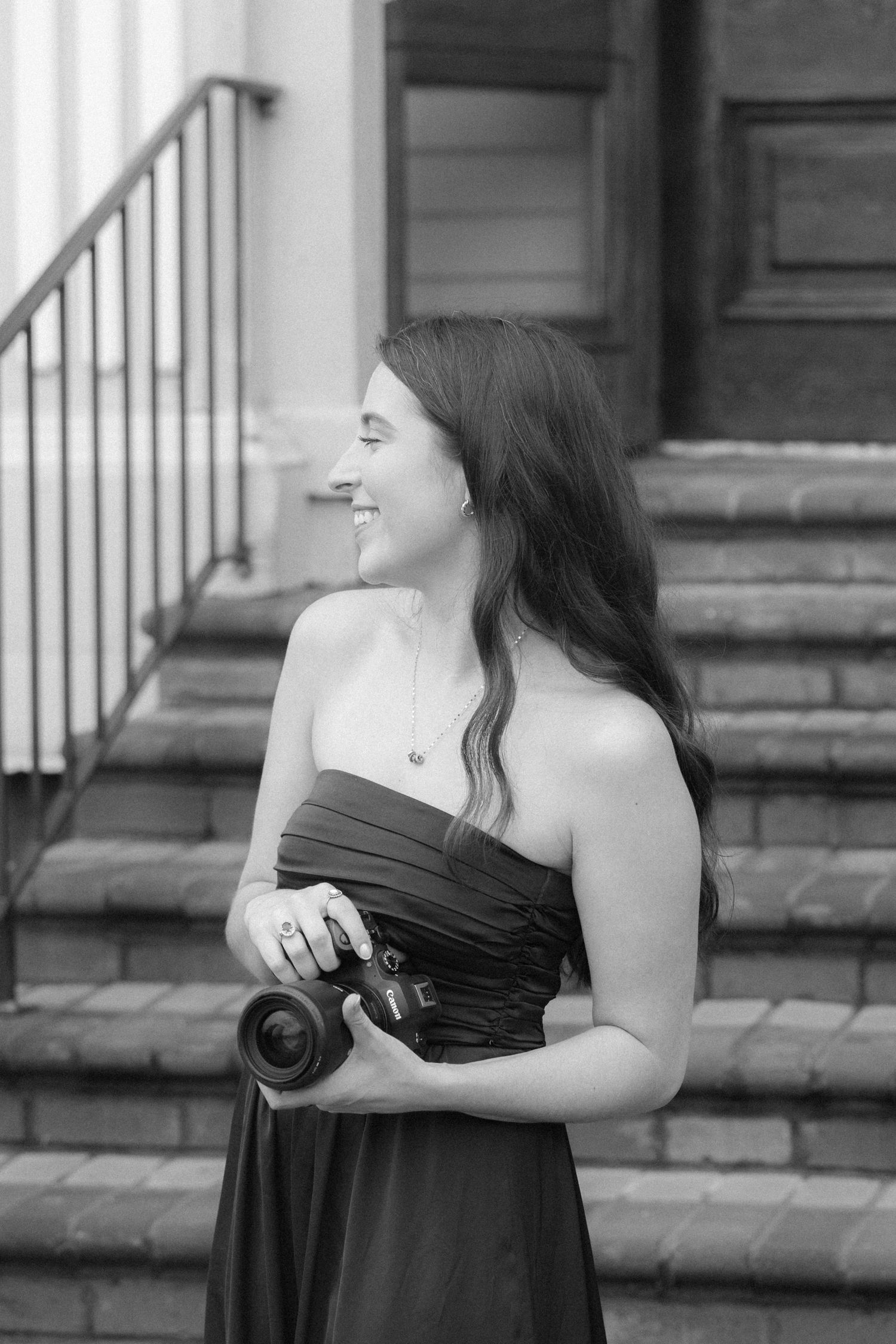 Smiling woman in strapless dress holding a camera on outdoor steps in black and white photo.