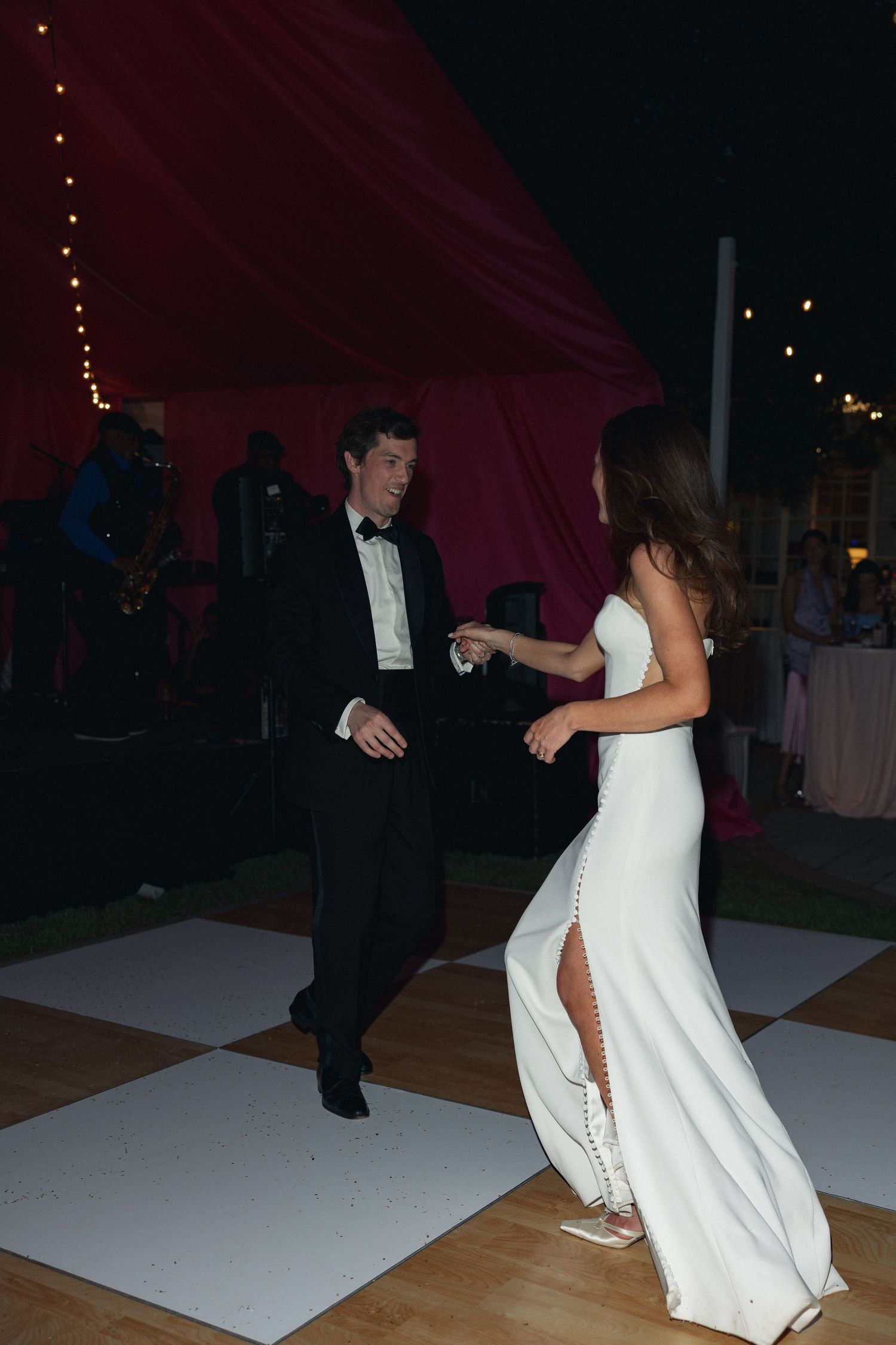 Bride in white gown and groom in tuxedo share first dance on outdoor floor at elegant evening wedding reception.