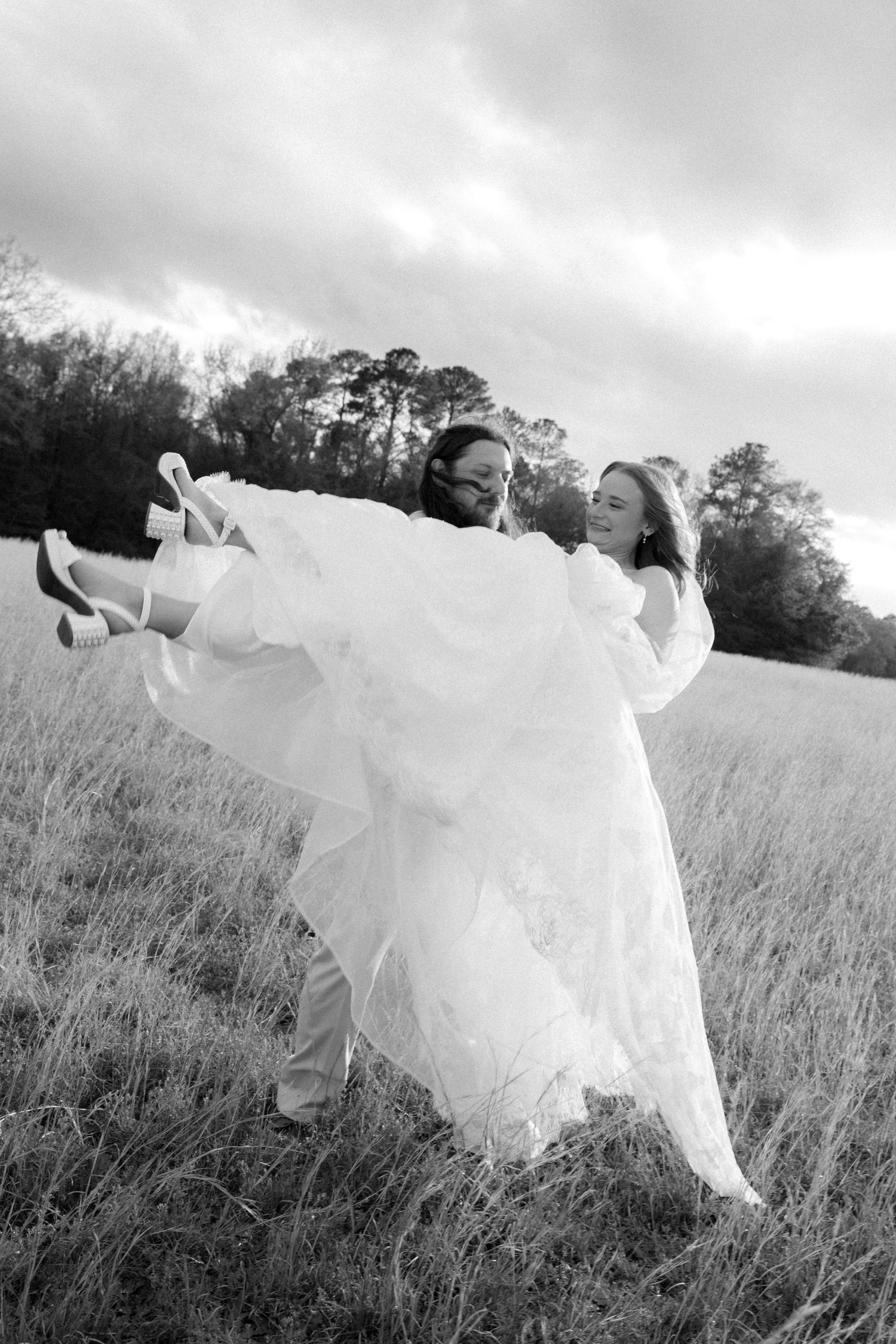 Black and white photo of groom swinging bride in wedding dress in a field.