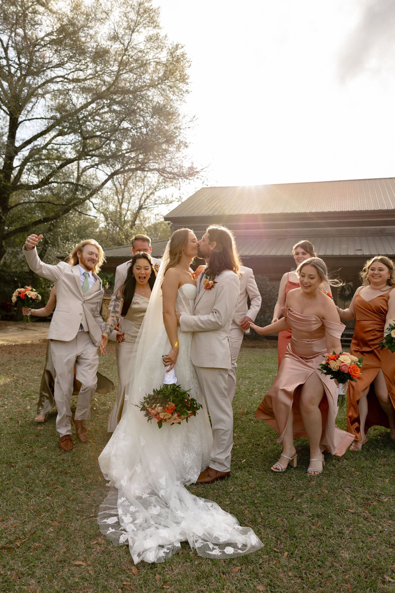 Bride and groom share a kiss surrounded by cheering wedding party in neutral and earth-tone attire outdoors.