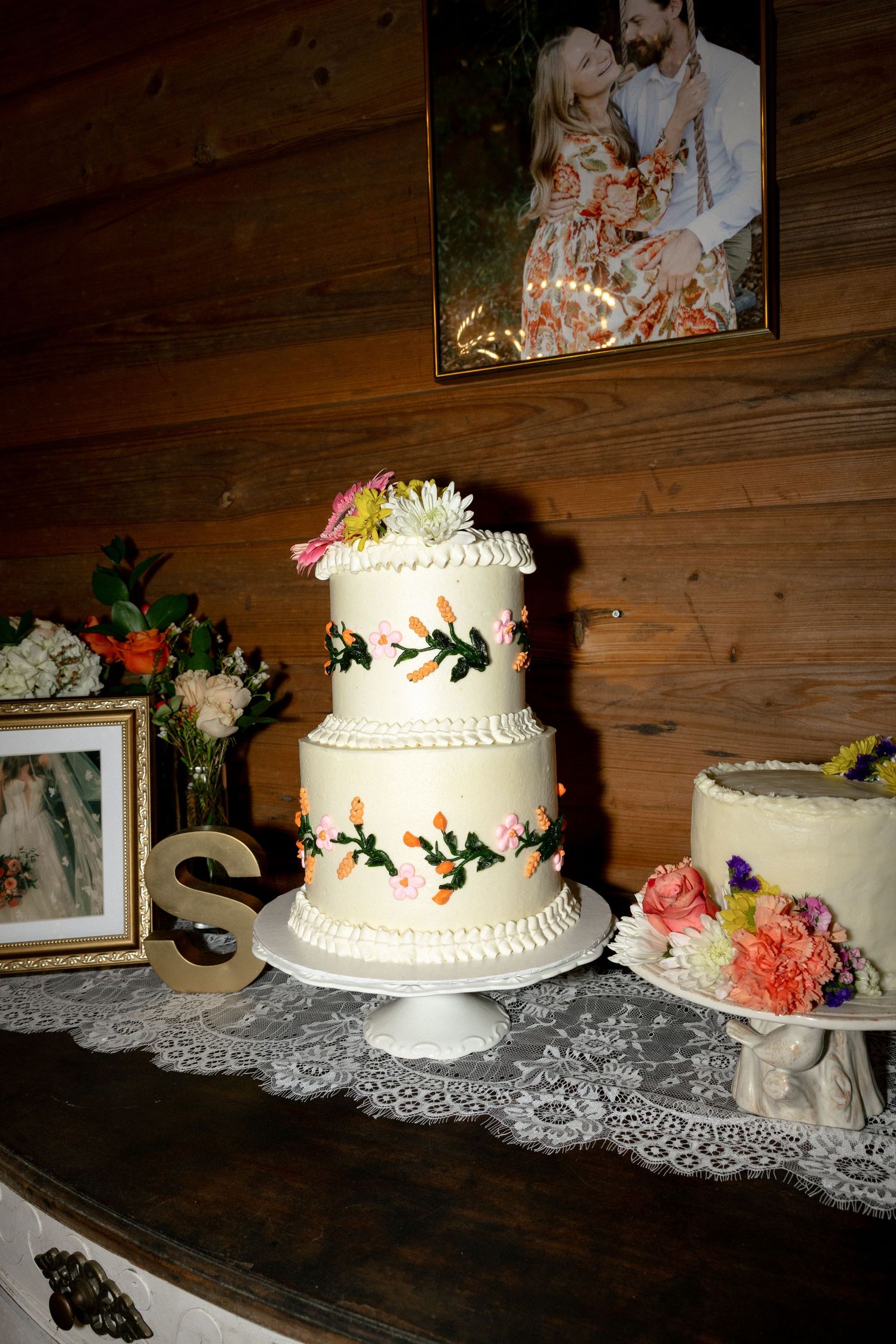 Two-tier white wedding cake with floral decorations displayed on lace-covered table in rustic barn.