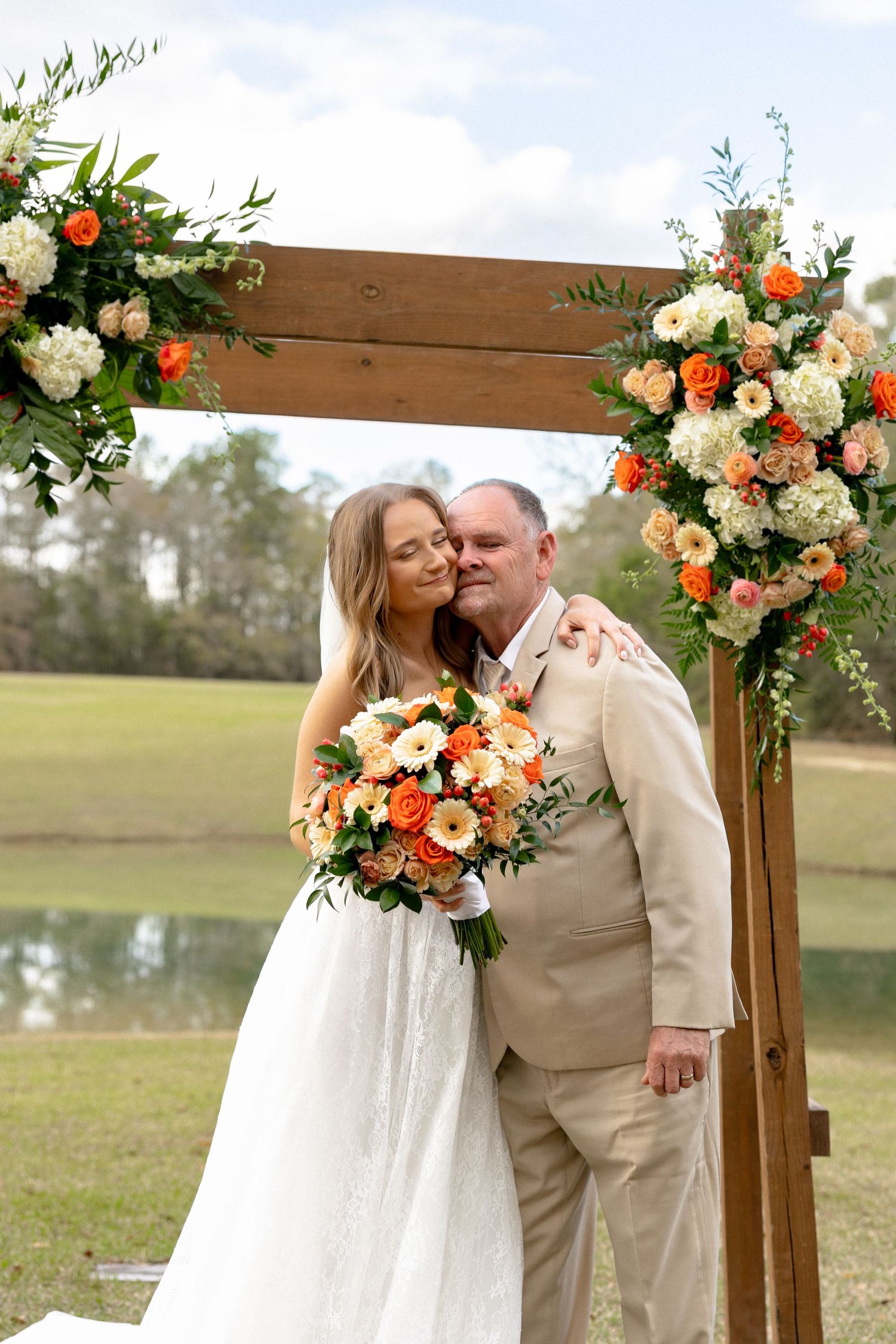 Bride and groom share tender moment under floral wedding arch with orange blooms.