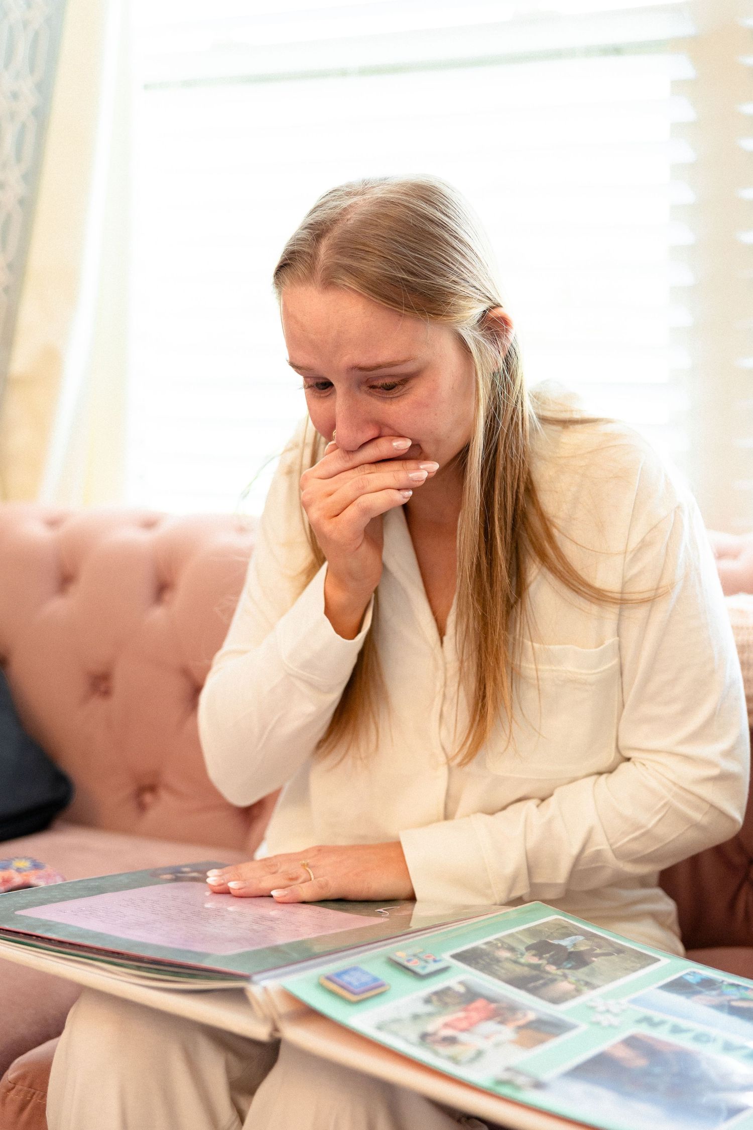 Tearful bride in cream pajamas looking at photo album on pink sofa before wedding.