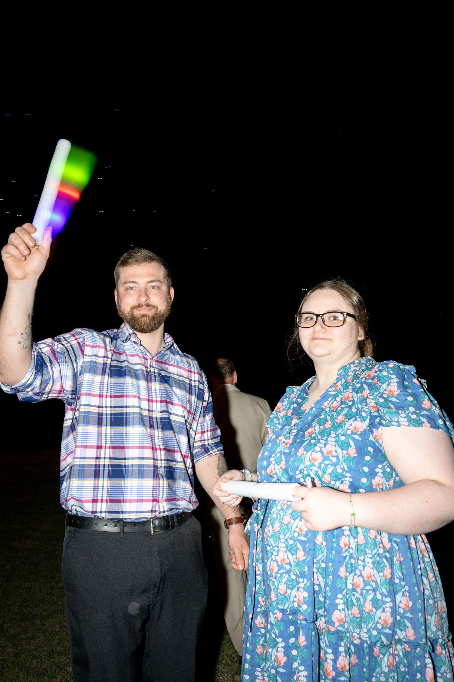 A man and woman holding glowing LED foam sticks at an outdoor nighttime event, smiling for the camera.