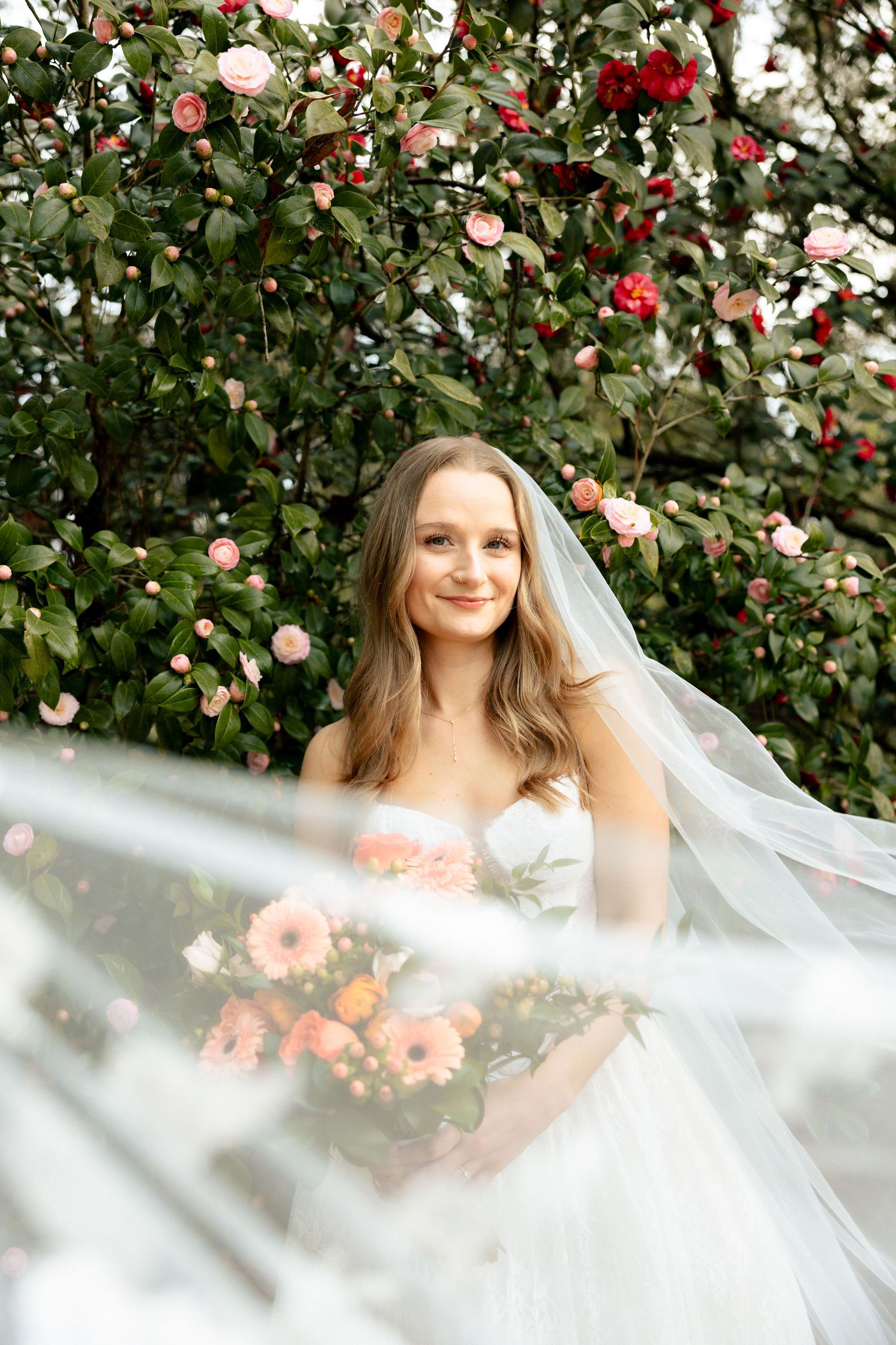 Smiling bride in strapless white gown and veil holds pink bouquet before blooming camellia flower tree.