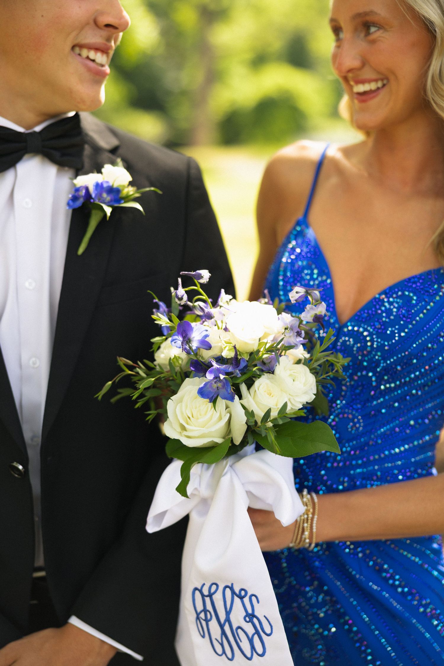 Smiling prom couple holding blue and white floral bouquet with monogrammed ribbon outdoors.