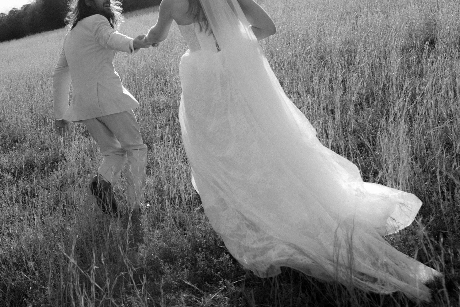 Black and white photo of bride and groom holding hands while walking through a grassy field.