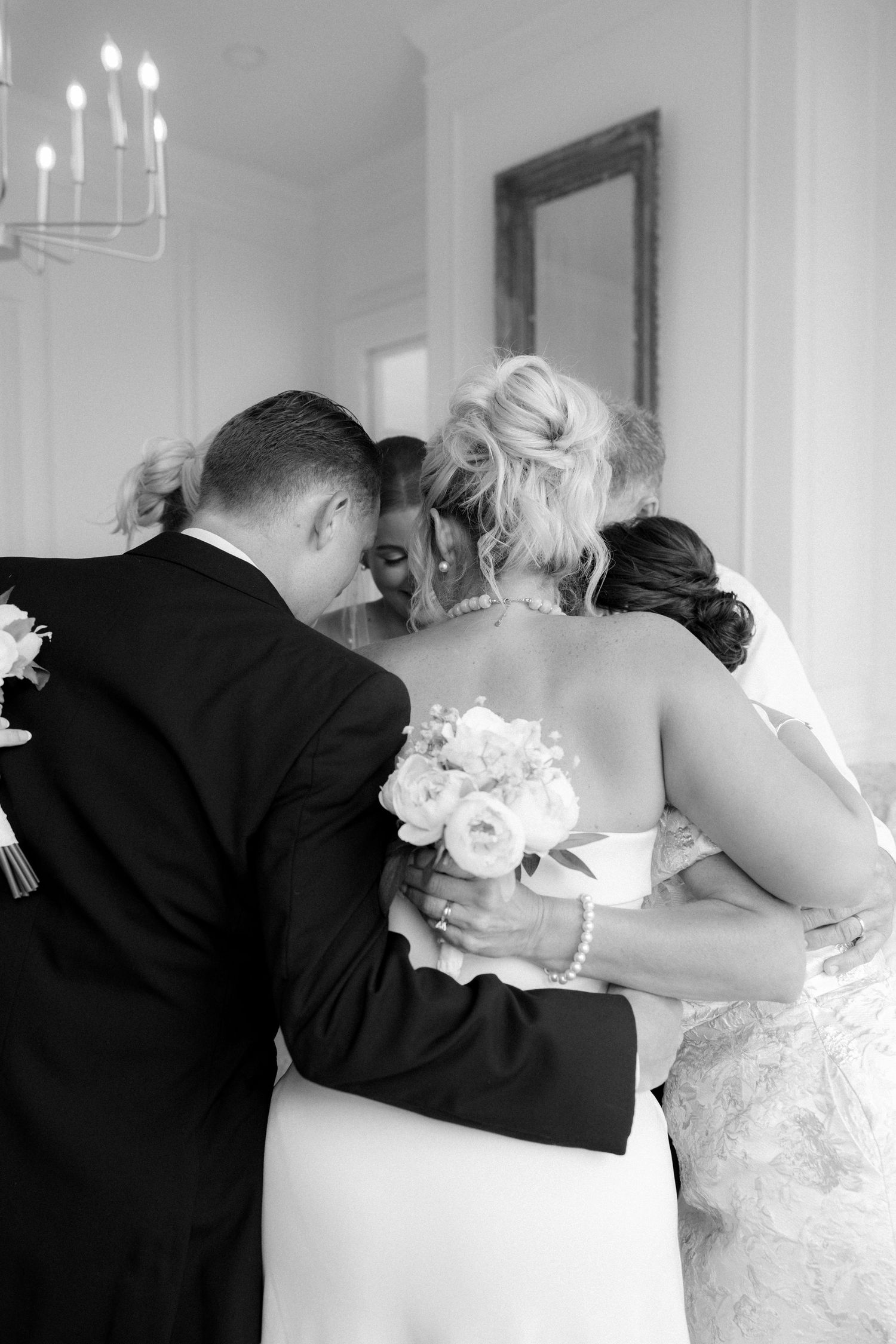 Black and white wedding photo of bride and groom embracing with guests, bride holding bouquet, reflected in mirror.