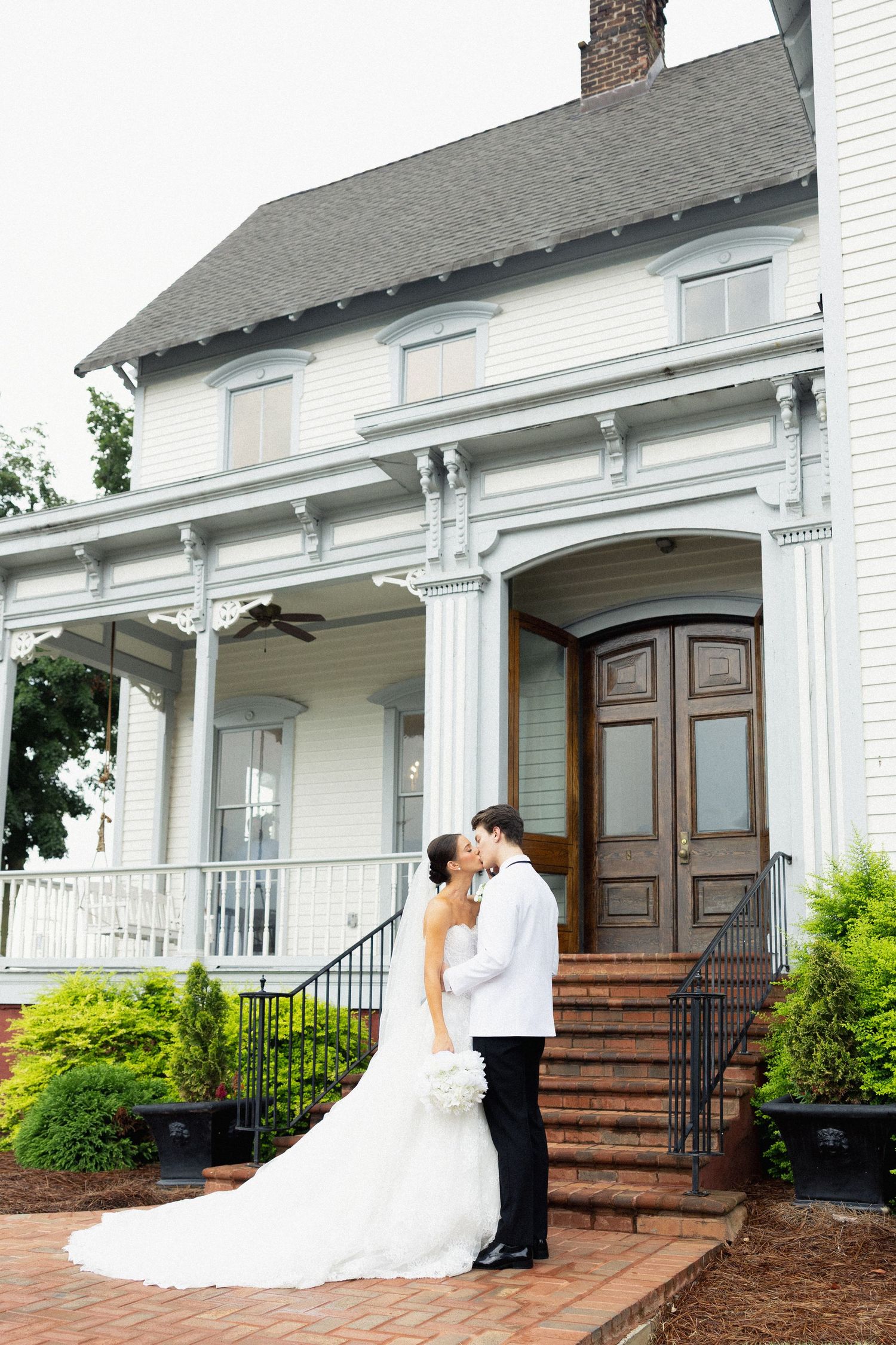 Bride and groom share a kiss in front of a white Victorian mansion with brick steps and elegant columns.