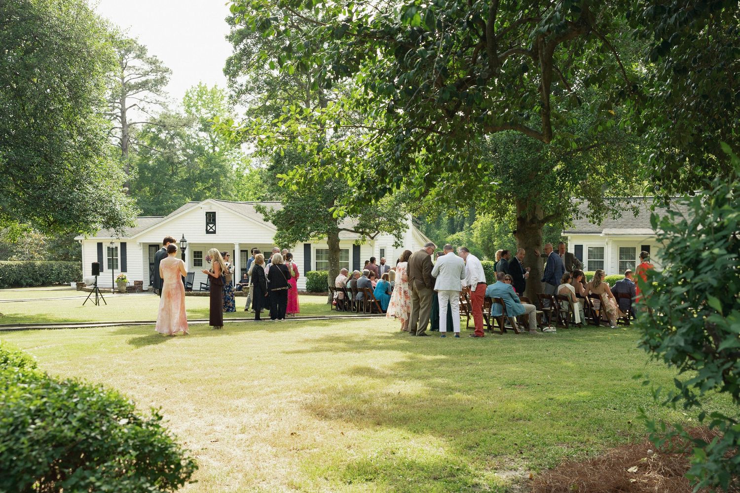 Outdoor wedding ceremony on a green lawn with guests gathered near a white house surrounded by tall trees.