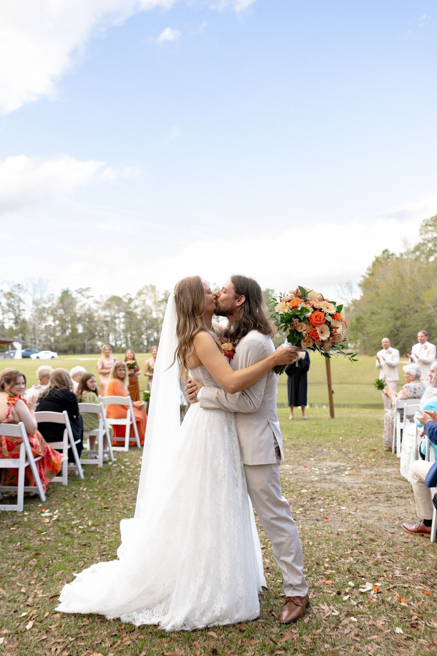Newlywed couple shares first kiss at outdoor wedding ceremony as guests cheer with orange floral bouquet.