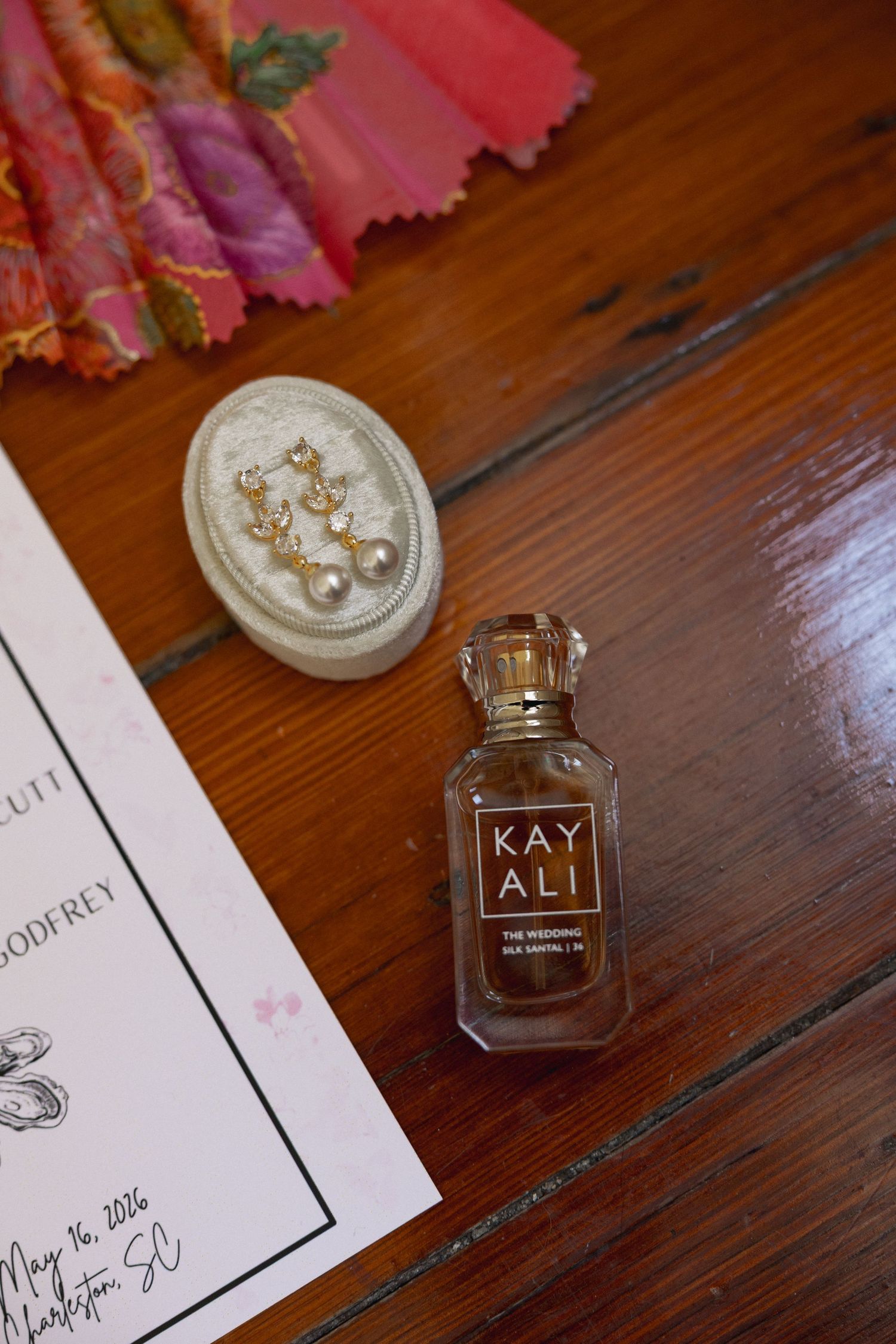 Kayali perfume bottle beside pearl drop earrings on velvet tray with wedding invitation on wooden surface.