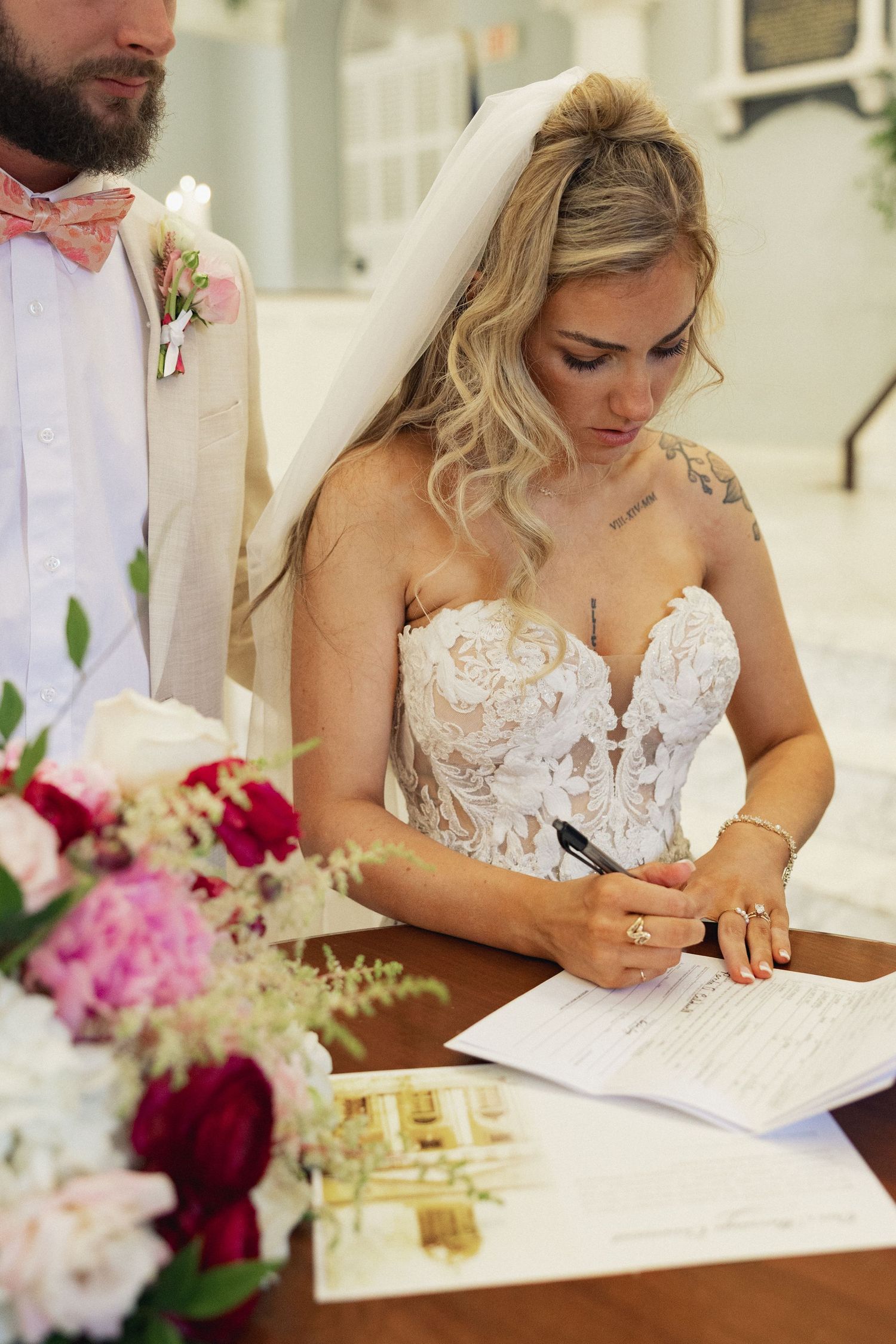 Bride in lace strapless gown and veil signing marriage certificate at wooden table with pink floral arrangement.