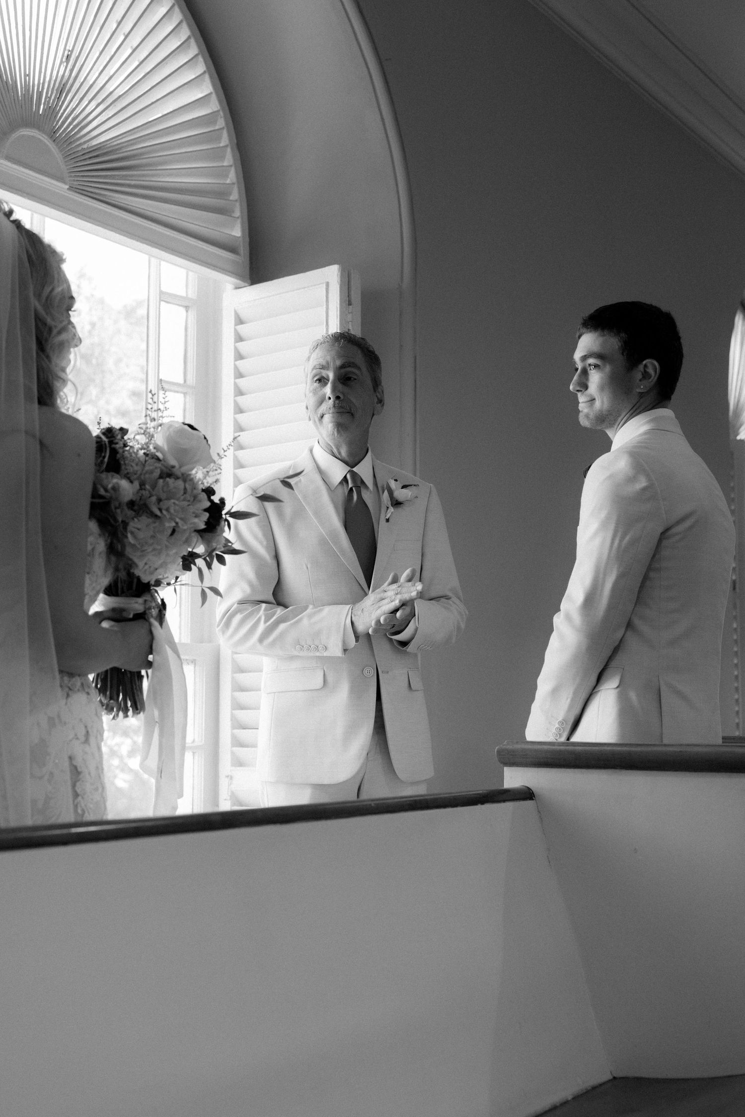 Two men in white suits stand in an elegant arched doorway, one holding a bridal bouquet at a wedding ceremony.