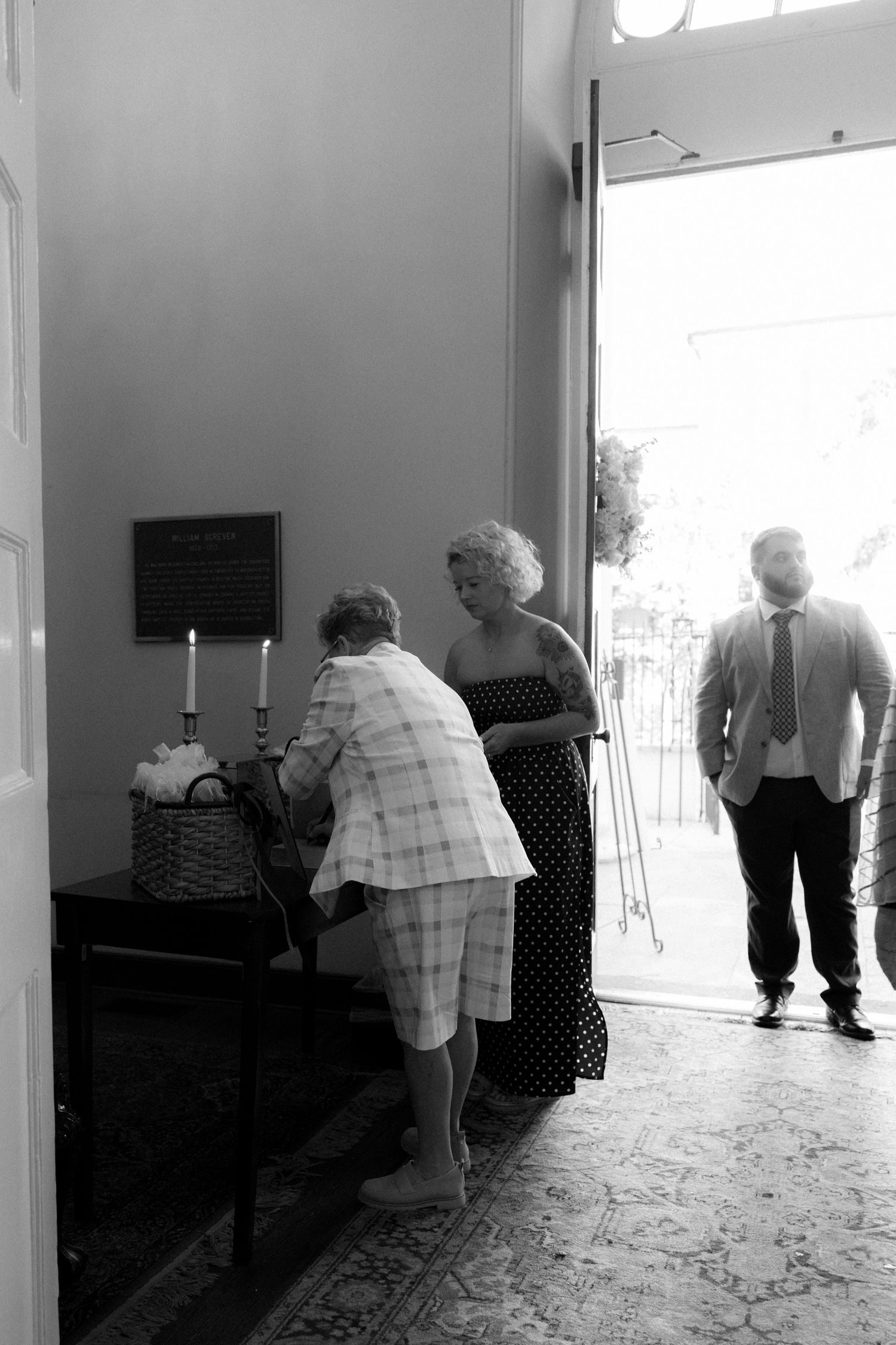 Black and white photo of wedding guests arriving at a church entrance, with a woman signing in at a table.