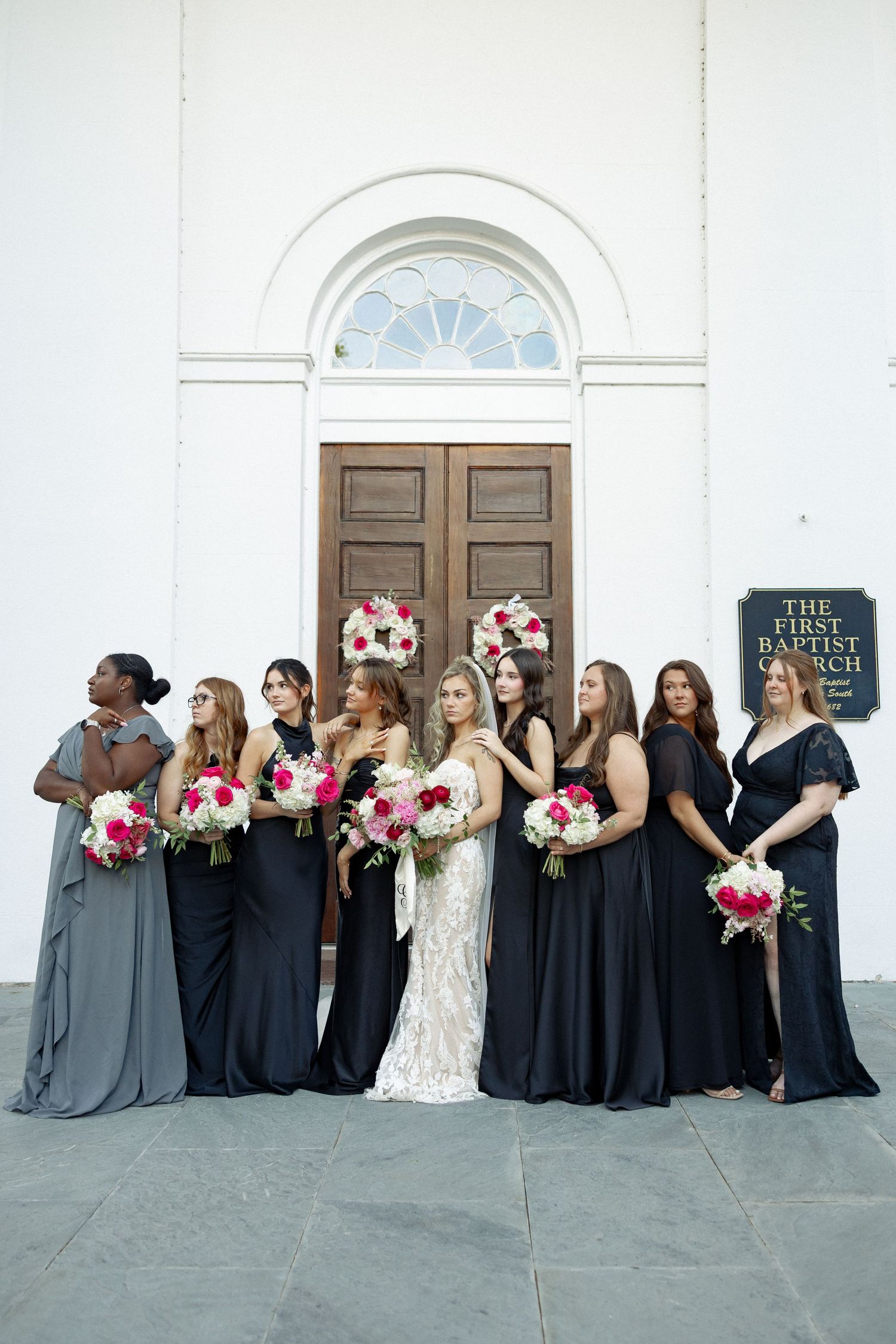 Bride in lace gown with bridesmaids in navy and gray dresses holding pink bouquets outside First Baptist Church.