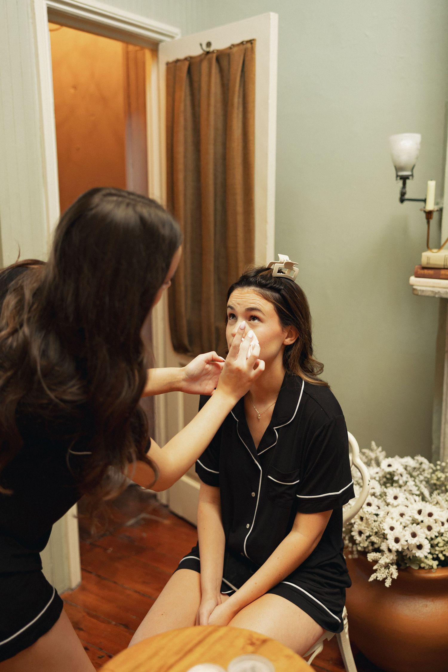 A bridesmaid applies makeup to a seated woman in black pajamas near a mirror with white flowers nearby.