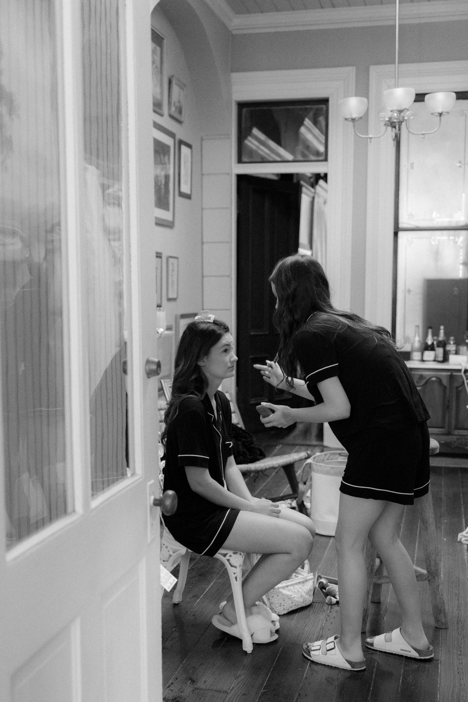 Two bridesmaids in matching black dresses chat and laugh together in a hallway during wedding preparations.