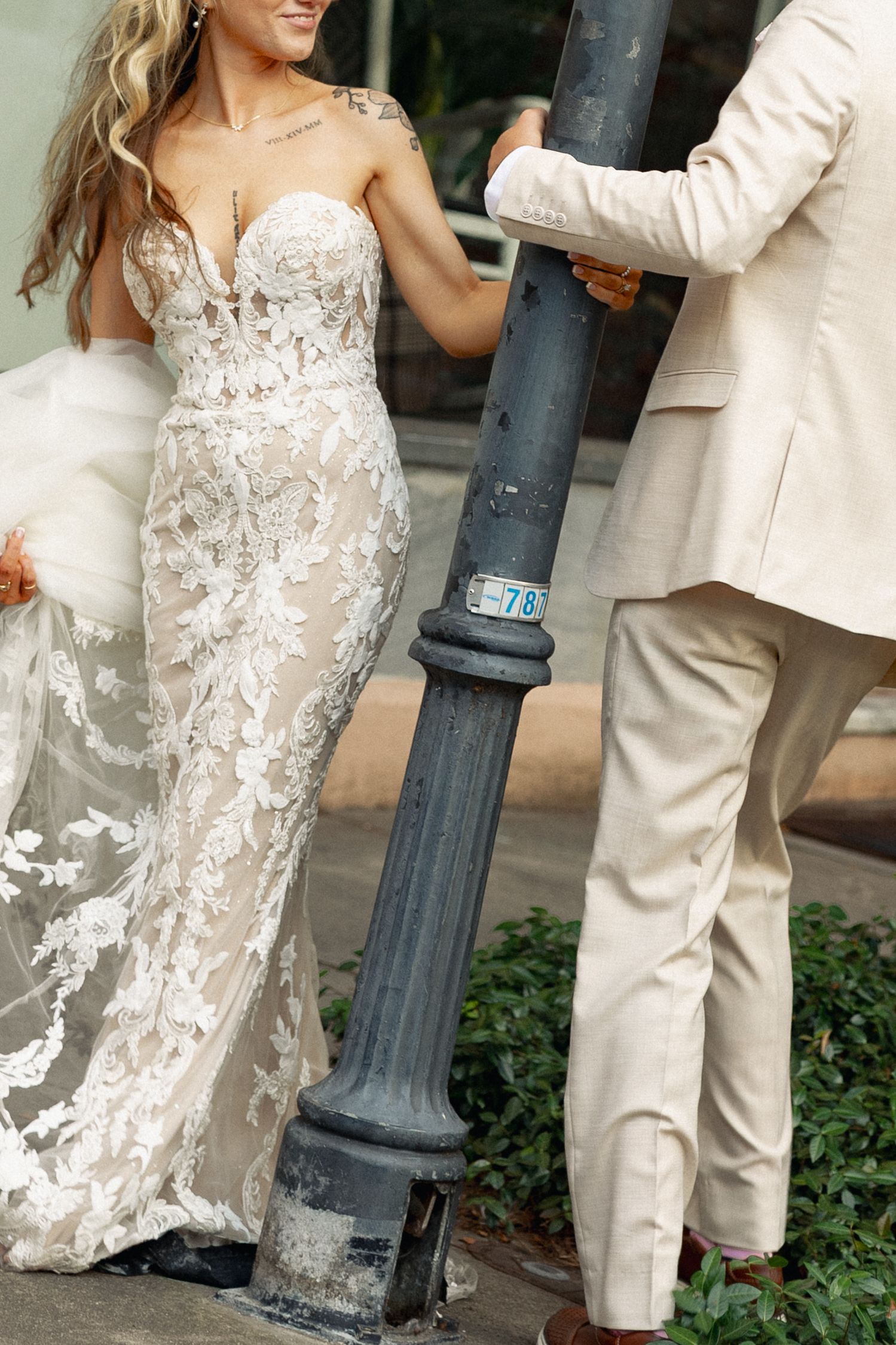 Bride in strapless lace wedding gown and groom in beige suit pose together beside a vintage street lamp.