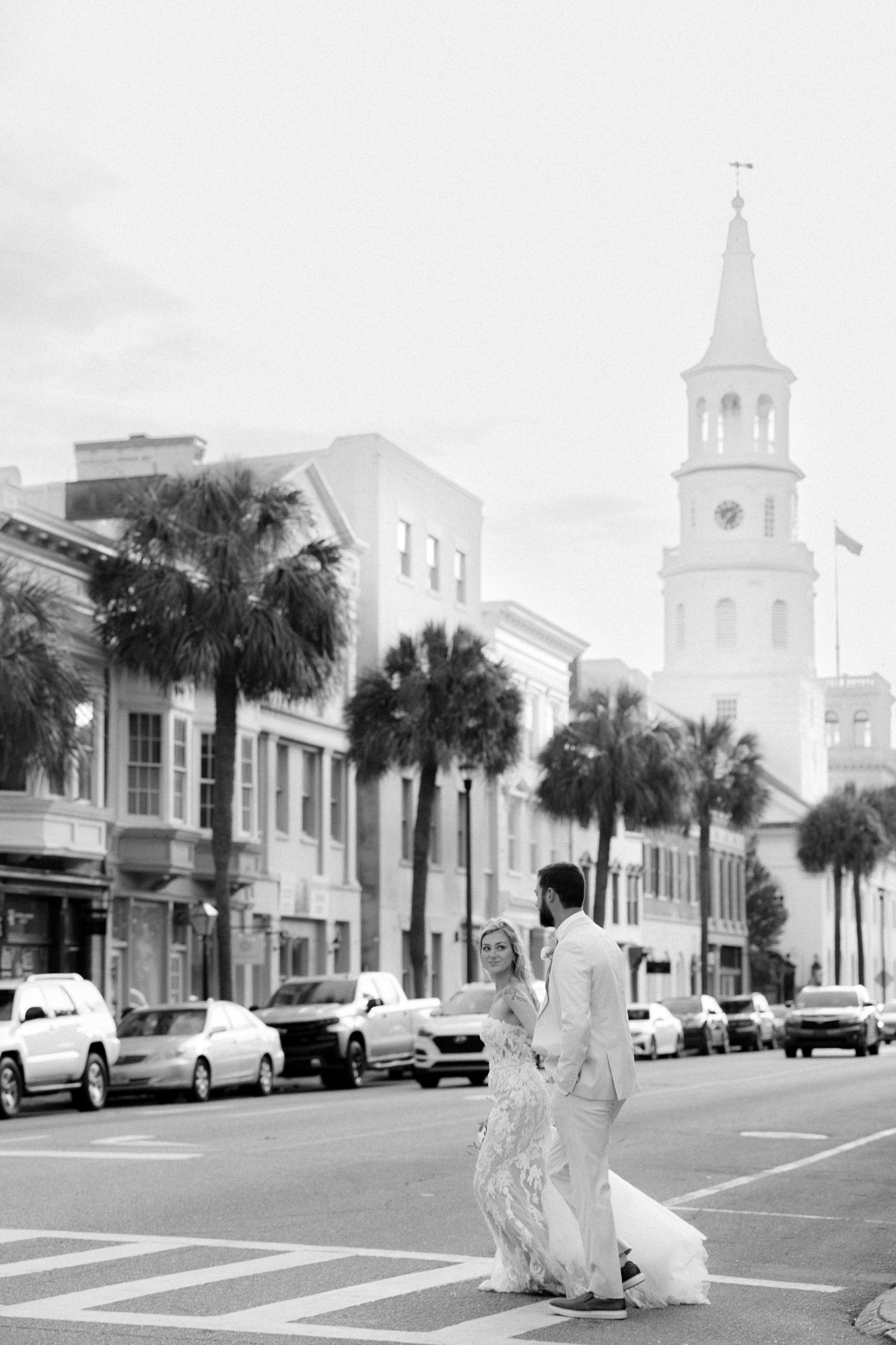 Black and white photo of bride and groom crossing street in Charleston with palm trees and church steeple in background.