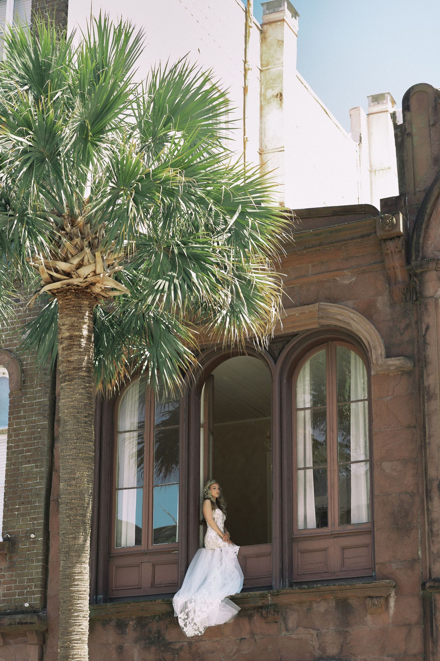 Bride in flowing white wedding dress standing in arched window of historic brick building with palm tree.
