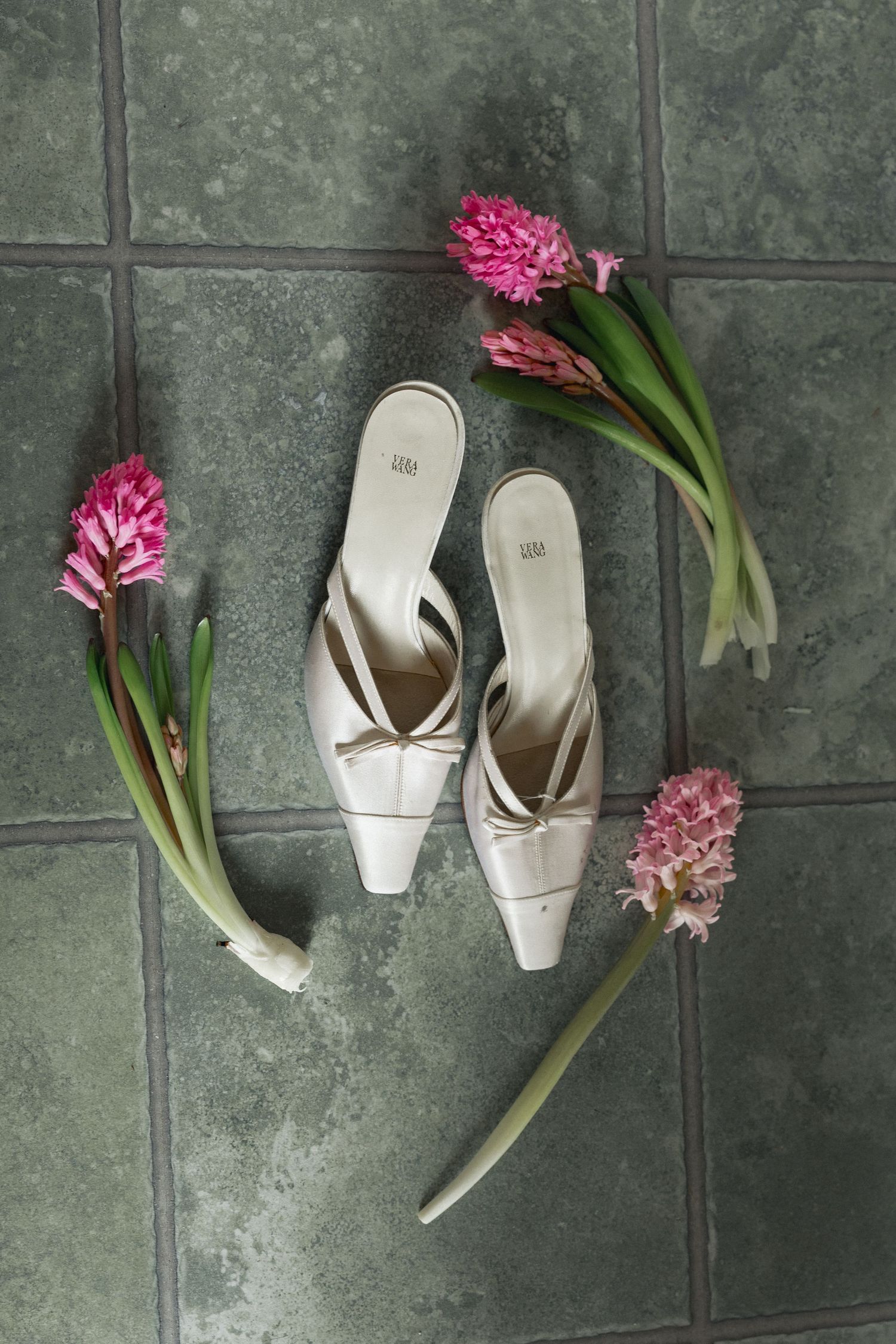 Elegant ivory bridal mule heels surrounded by pink hyacinth flowers on green tile floor.
