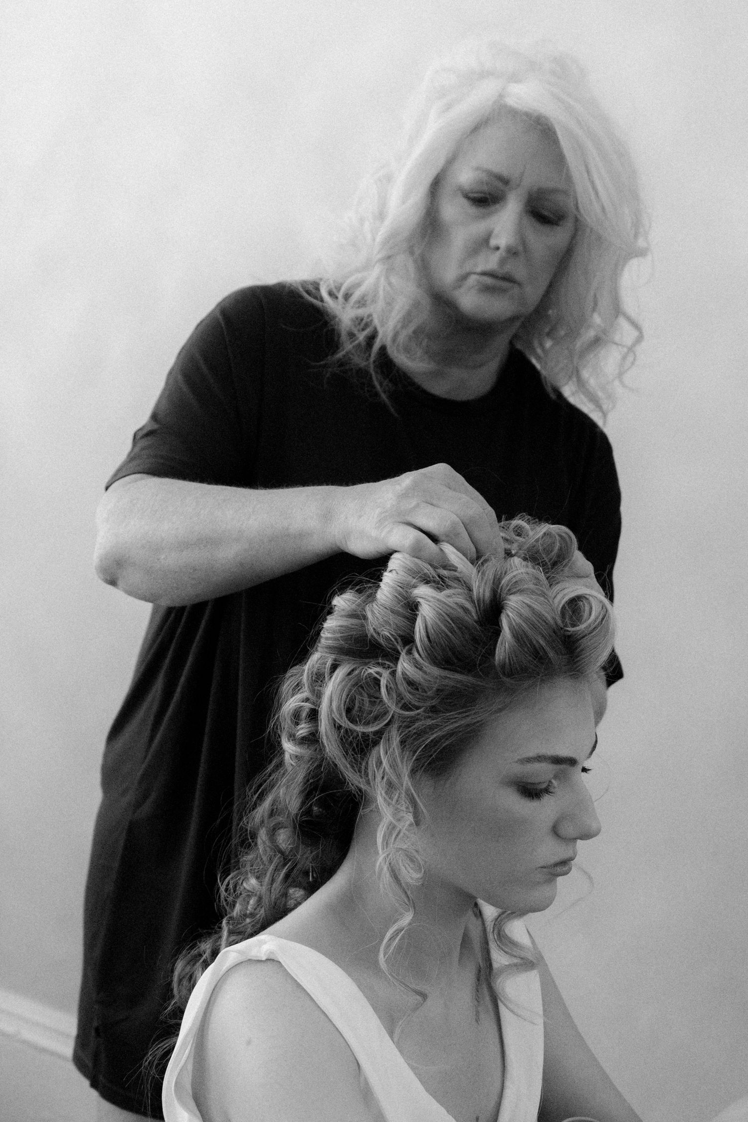 Black and white photo of a hairstylist pinning elegant updo curls on a bride in a salon.