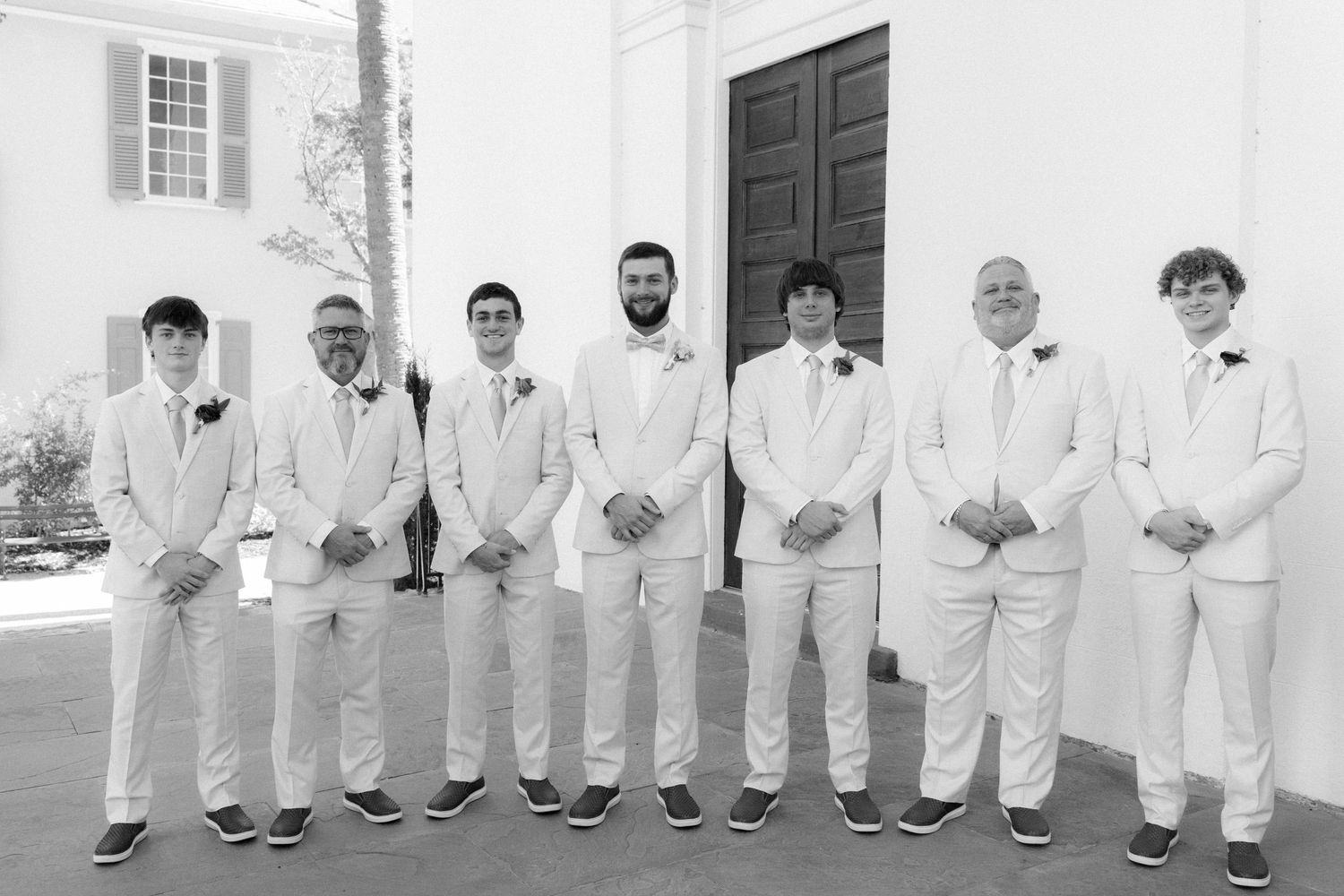 Groom and groomsmen posing in matching white suits outside a church in a classic black and white wedding photo.