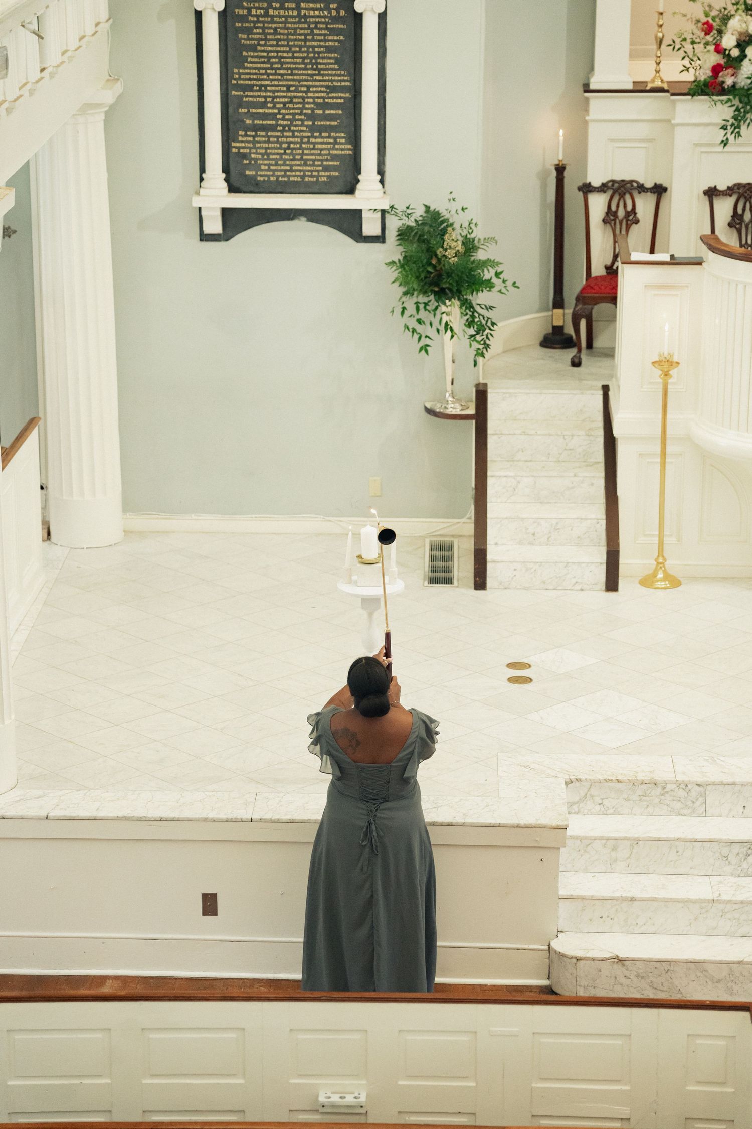 A woman in a gray gown lights candles at a church altar using a long candle lighter during a ceremony.