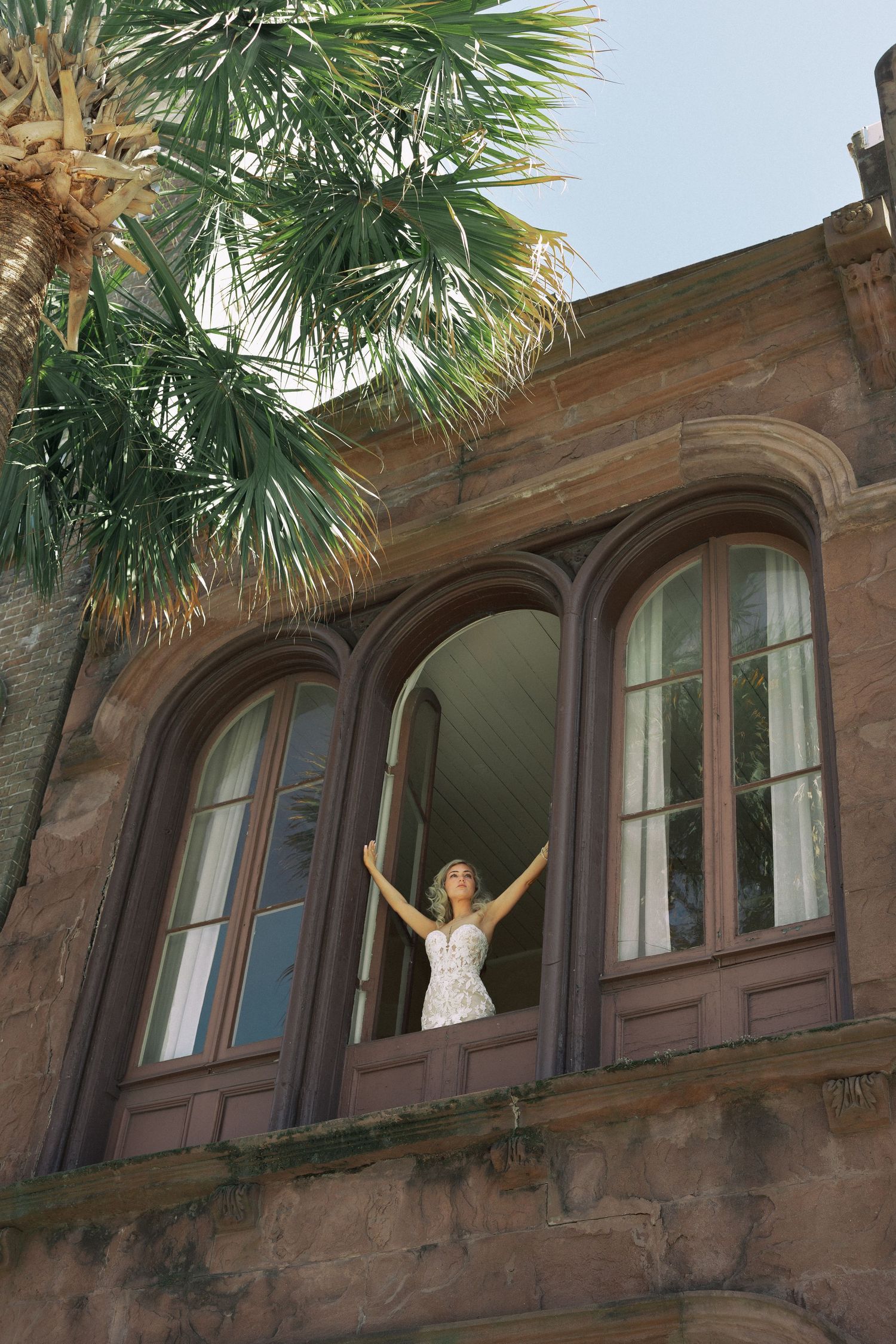 Bride in white dress standing in arched window of historic stone building with palm tree.