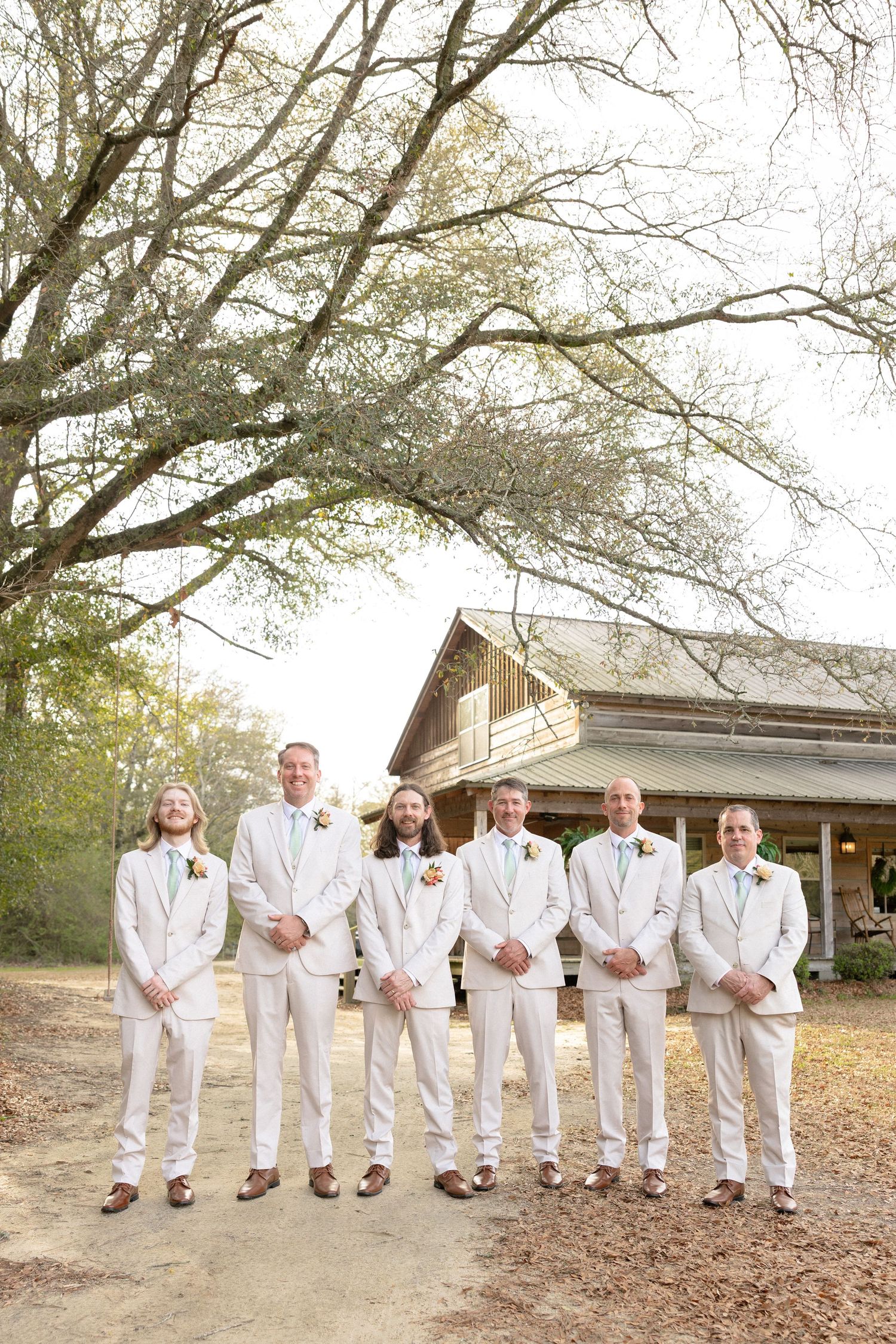Six groomsmen in matching cream suits with boutonnieres stand together outside a rustic barn venue.