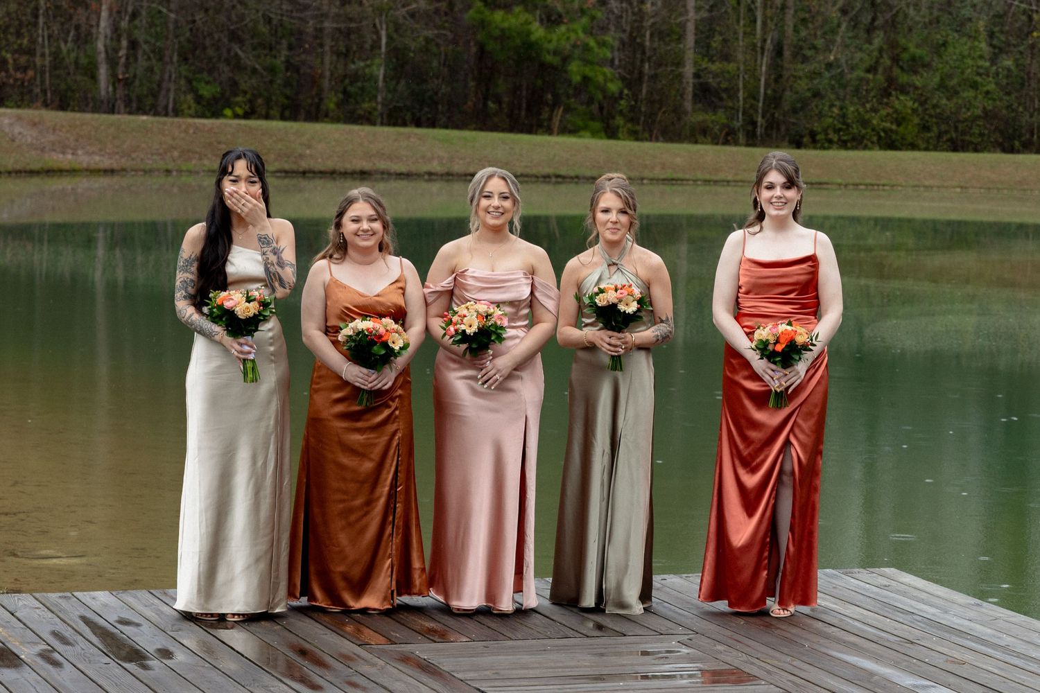 Five bridesmaids in mismatched earth-tone satin gowns holding orange bouquets pose on a wooden dock by a serene lake.