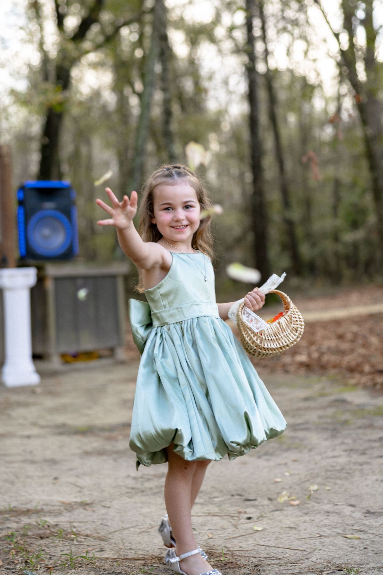 An adorable smiling flower girl in mint green dress waves while holding a wicker basket at an outdoor wedding ceremony.