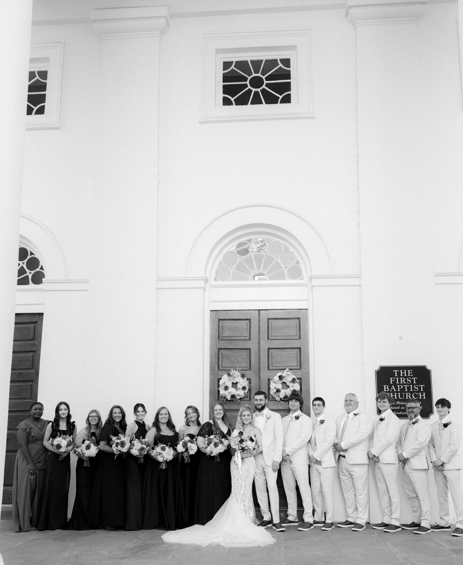 Black and white photo of wedding party posed outside First Baptist Church with groomsmen in light suits.