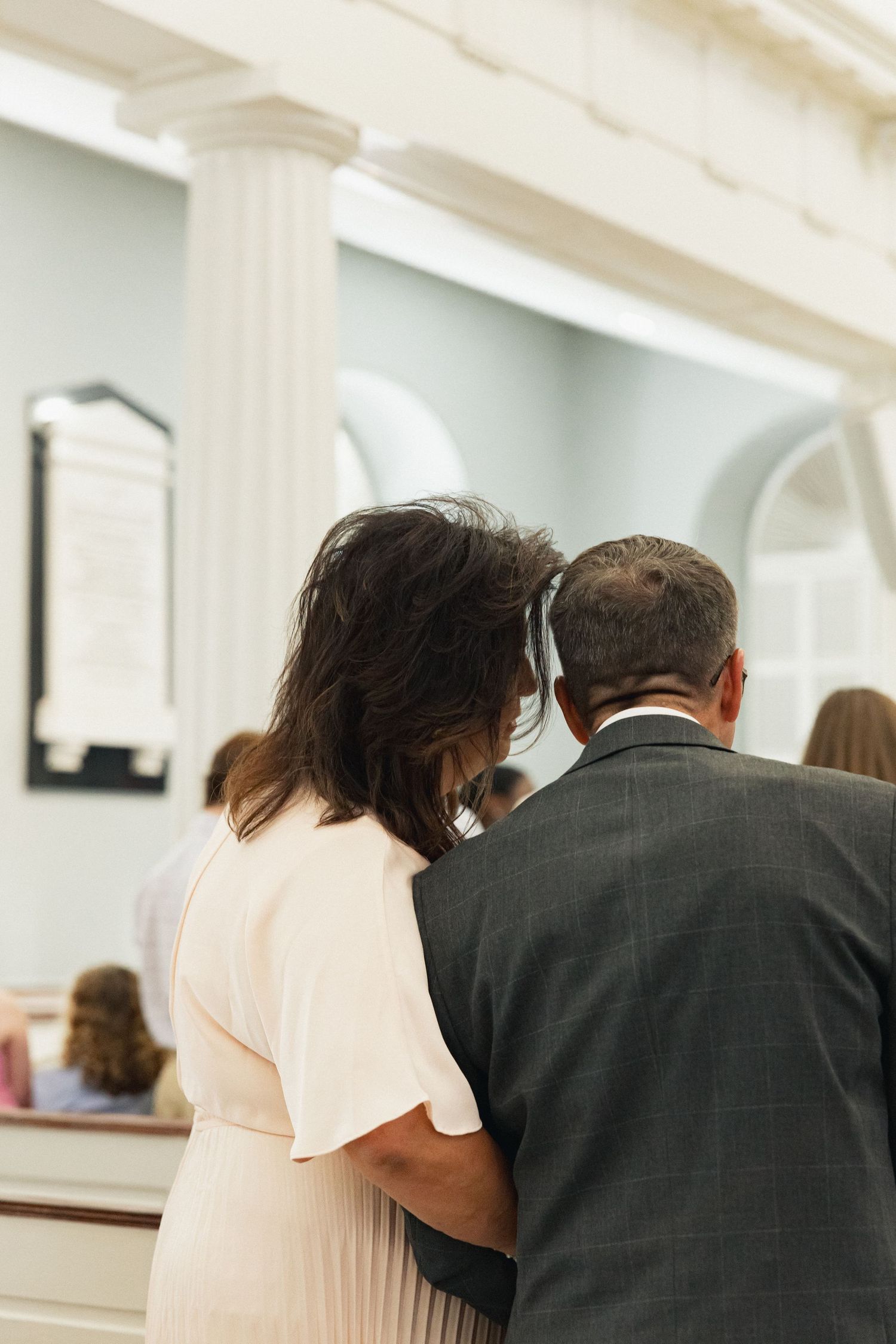 A couple stands arm in arm from behind, watching a wedding ceremony inside a church.