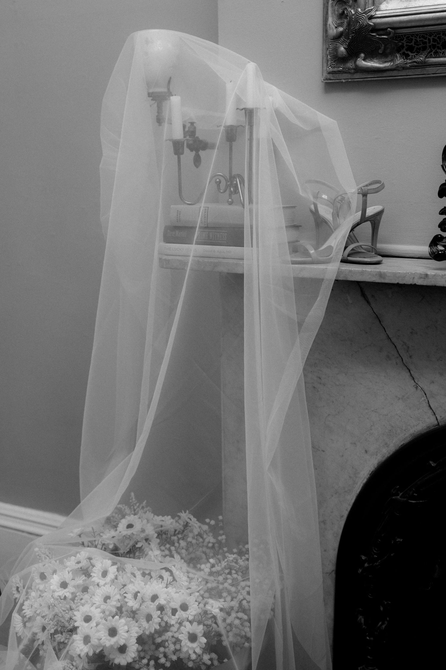 Black and white photo of a wedding veil draped over a chair near a fireplace with white daisy bouquet below.