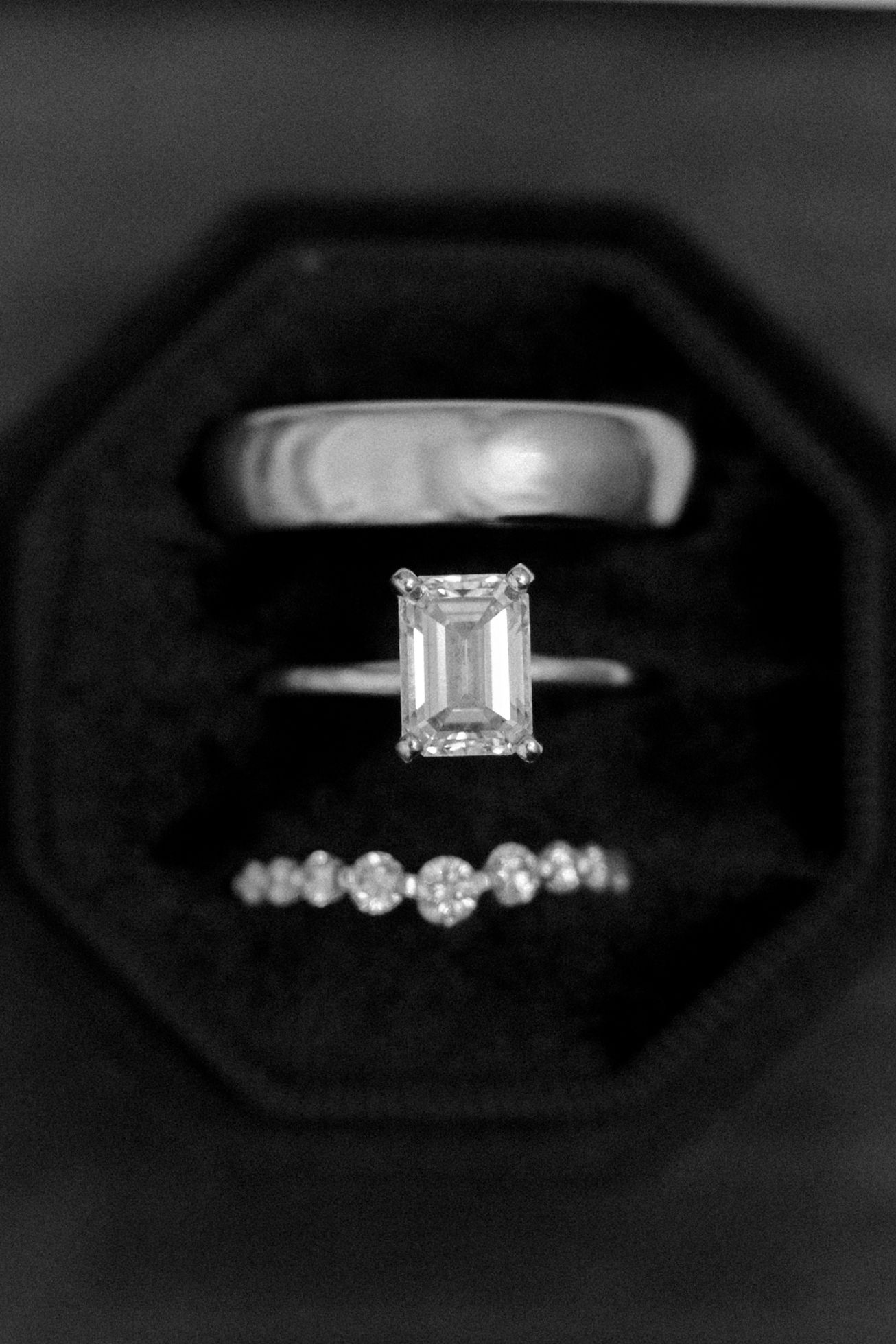 Black and white photo of emerald cut solitaire engagement ring with diamond band in black velvet octagon ring box.