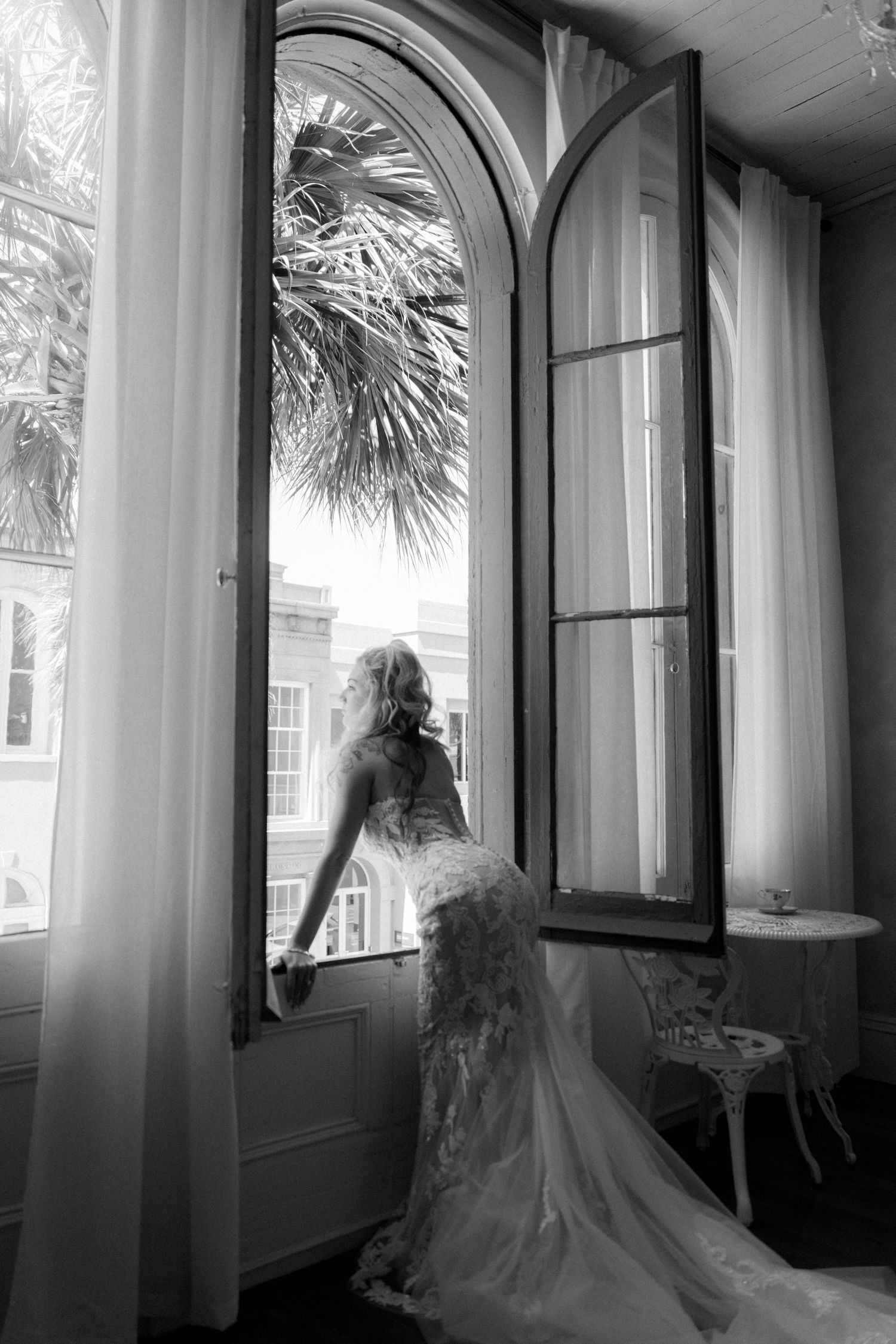 Black and white photo of bride in lace gown standing at open arched window with palm tree view.