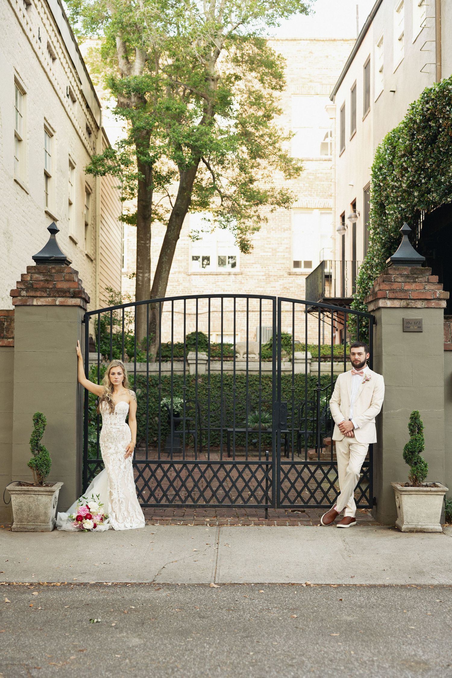 Bride and groom pose separately on either side of ornate black iron garden gate flanked by brick pillars.