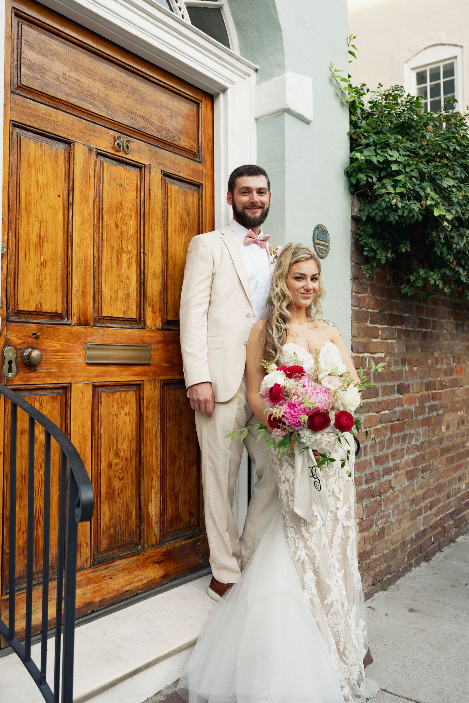 Bride and groom posing by a wooden door, she holds red and pink bouquet, he wears a tan suit with pink bow tie.
