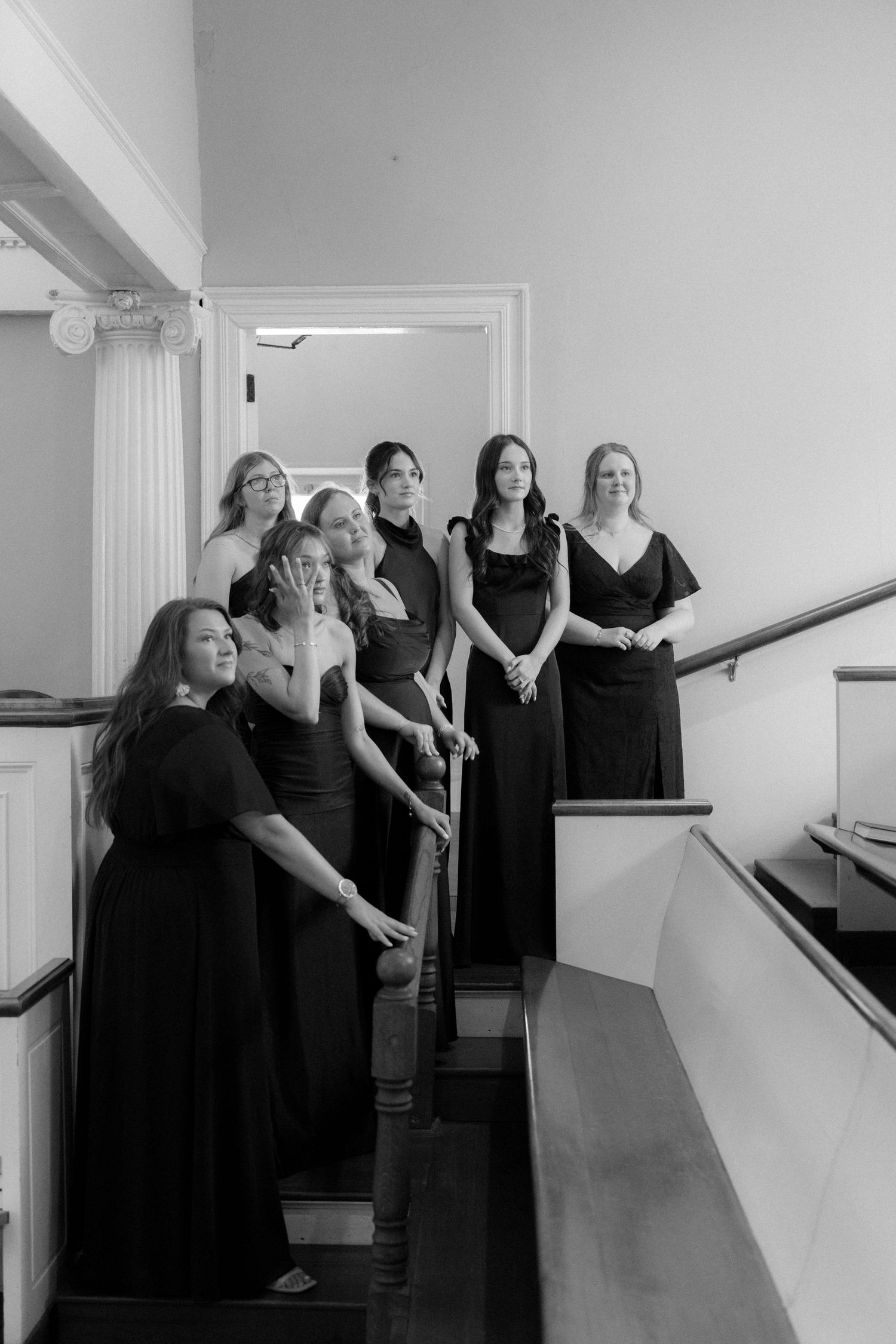 Black and white photo of seven bridesmaids in dark gowns posing on staircase at wedding venue.