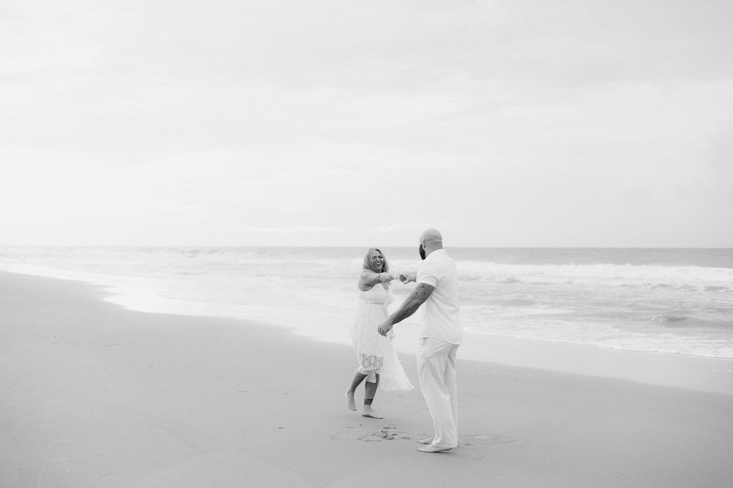 Couple dancing together on a sandy beach in black and white, waves crashing in the background.
