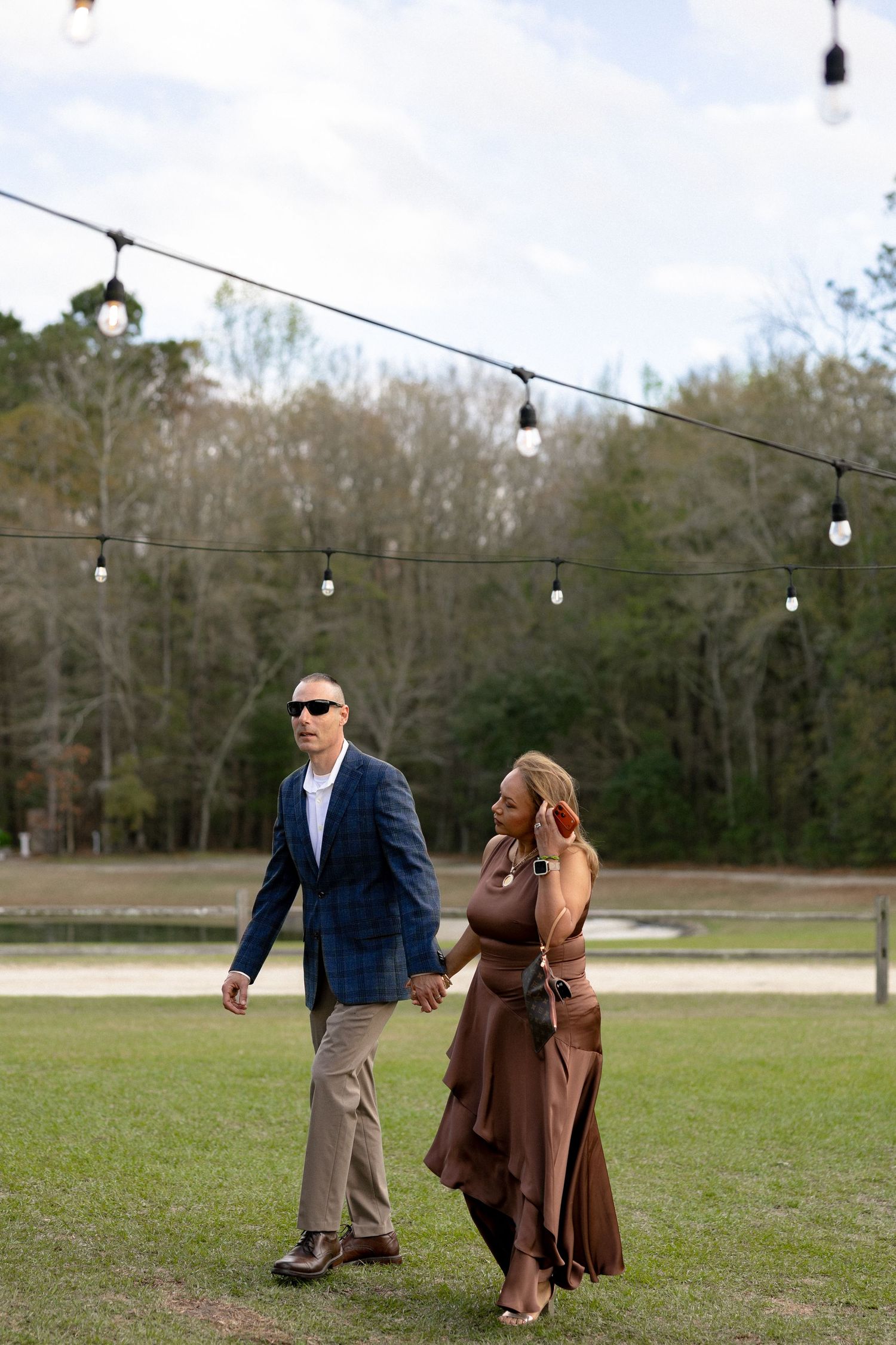 Elegant couple walking hand-in-hand on grass at an outdoor wedding venue with string lights.
